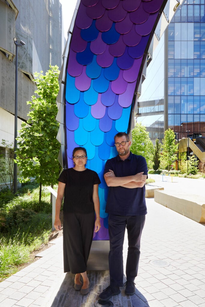 Two people stand outdoors beneath a large, curved, blue and purple installation with a scale-like pattern, surrounded by buildings and greenery.
