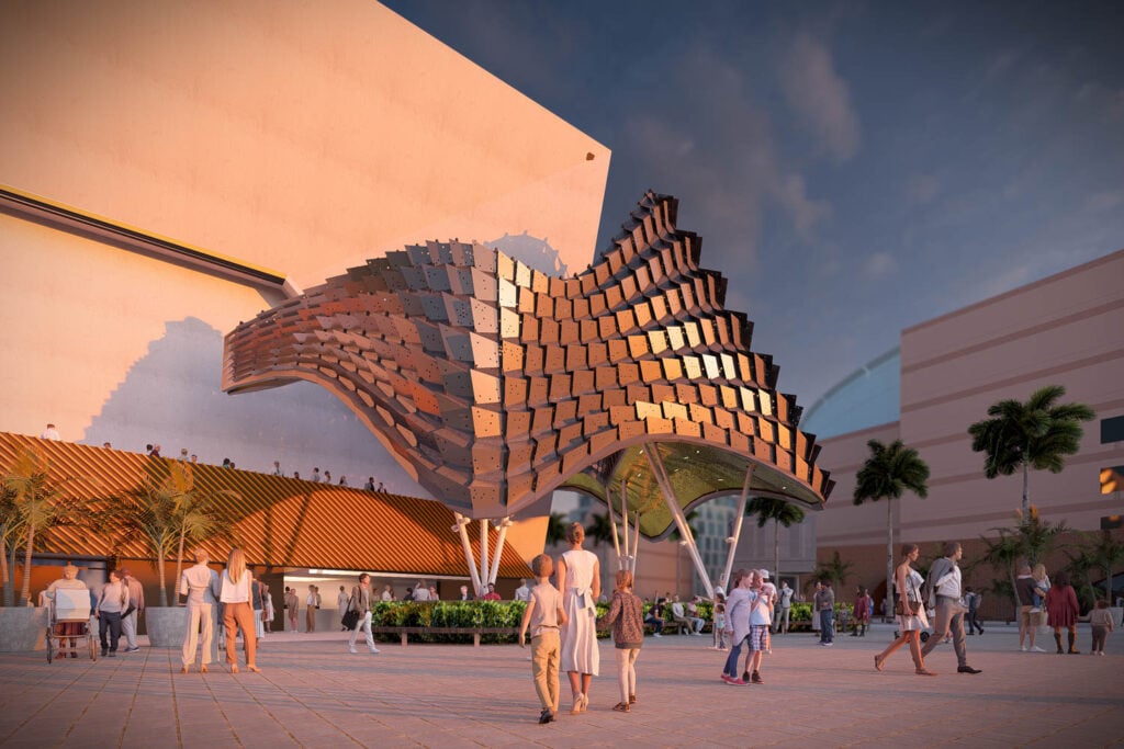 People walk and gather around a modern building with a large, curved, metallic architectural canopy, surrounded by palm trees at sunset.