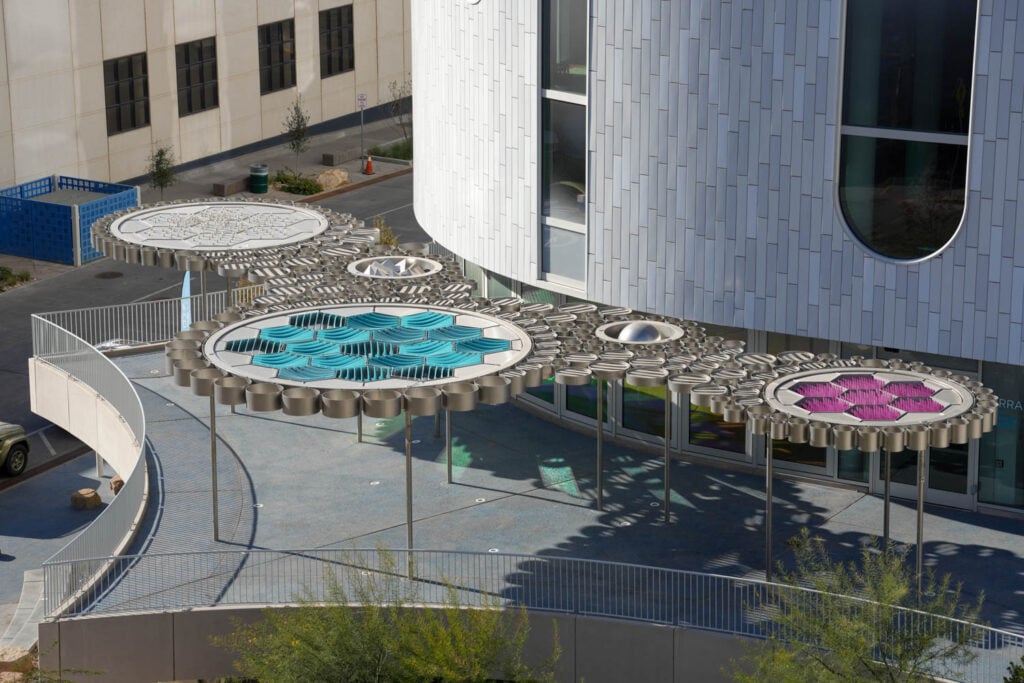 Metallic sculptural canopies with geometric patterns and colored panels provide shade outside a modern building with a curved, white facade.