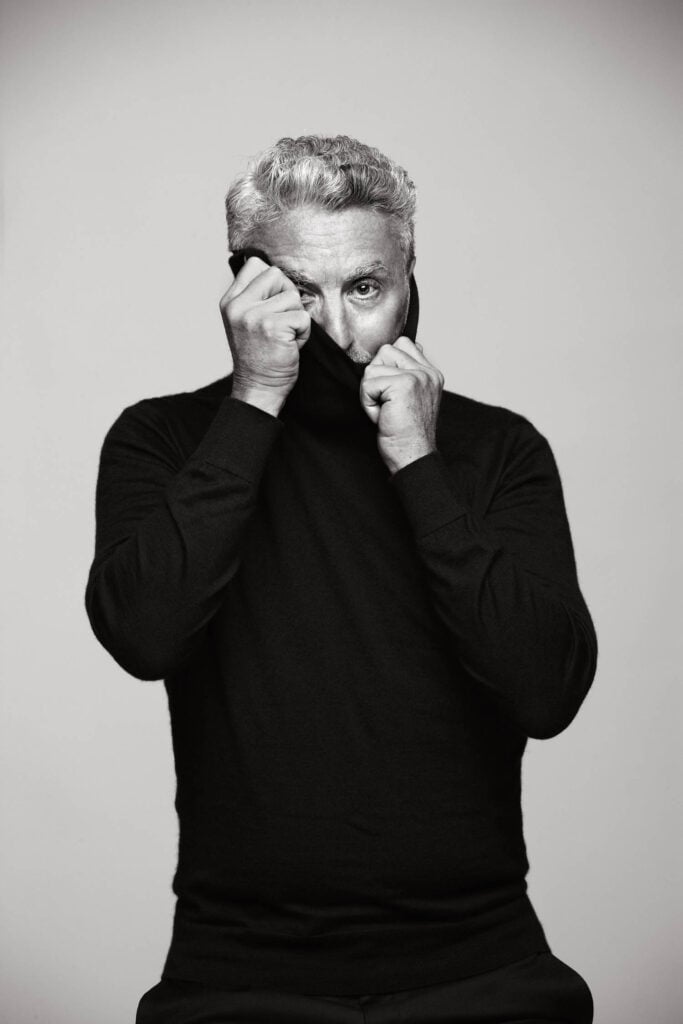 A man with gray hair, wearing a black long-sleeve shirt, pulls the collar up to partially cover his face; he stands against a plain, light background, echoing the understated style often seen in Grupo Habita’s modern spaces.