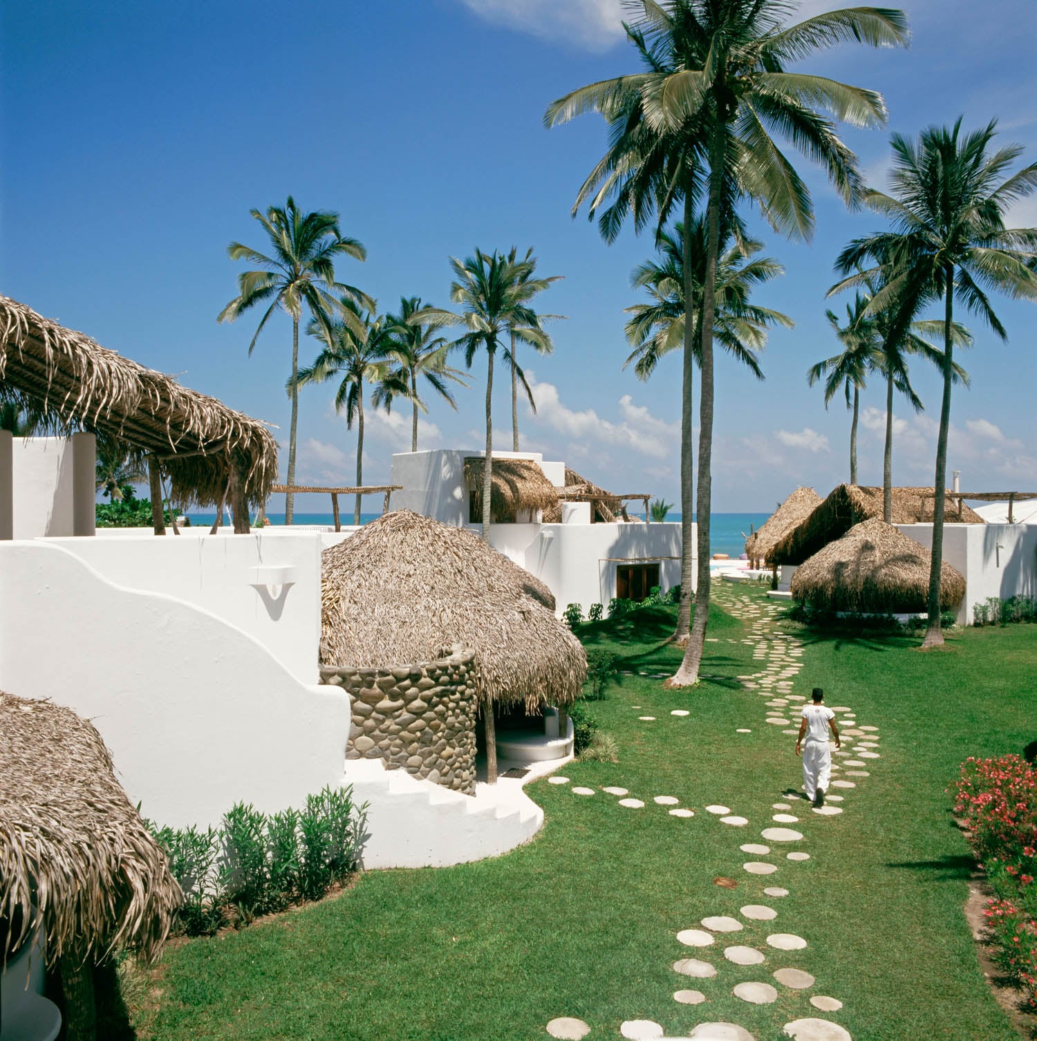 A person walks along a stone path between white, thatched-roof buildings and palm trees on a sunny day near the ocean.