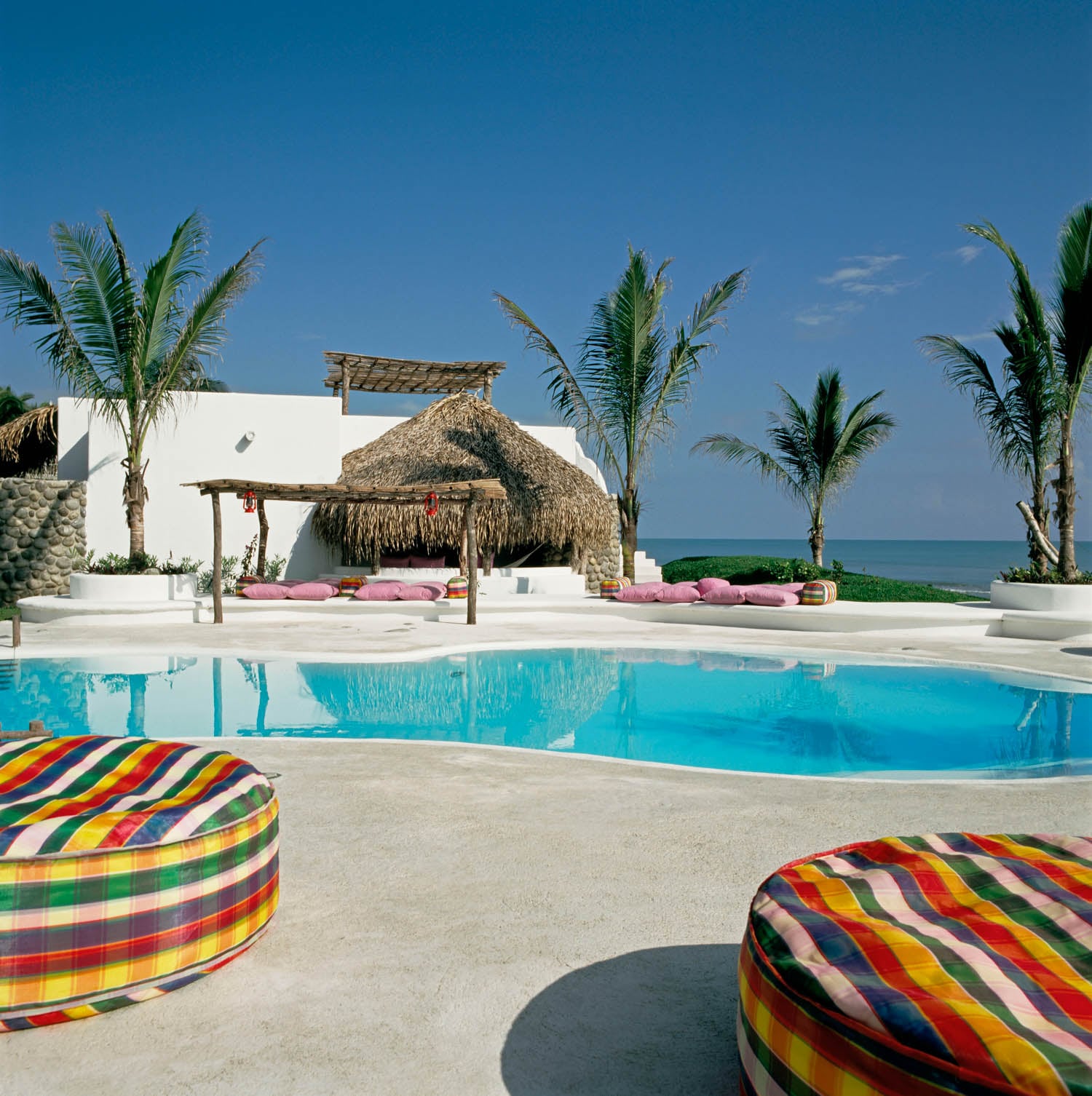 A tropical resort pool with clear blue water, palm trees, a thatched-roof hut, white walls, and colorful round loungers under a sunny sky with ocean visible in the background.