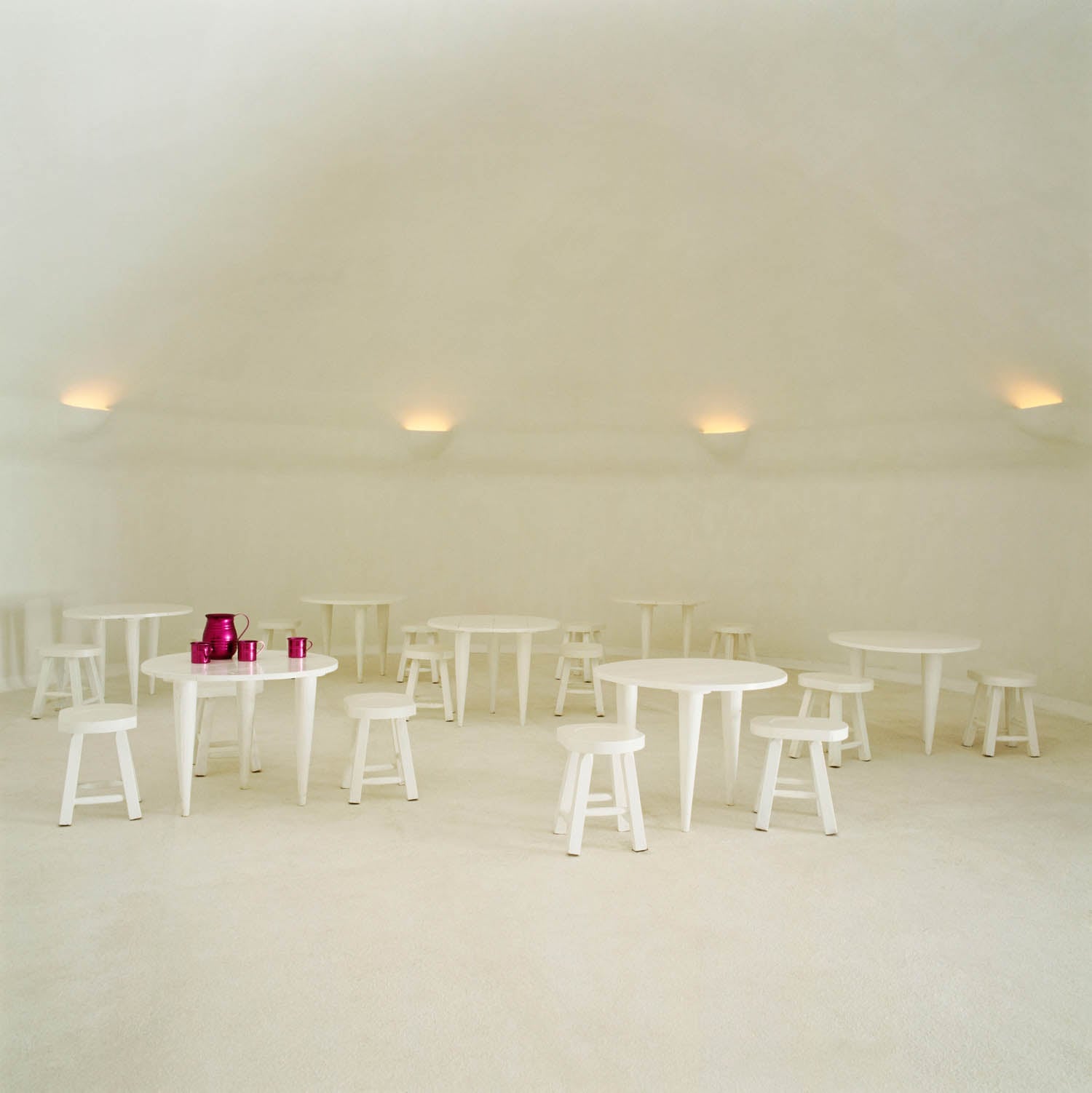 Minimalist room with multiple white round tables and stools, soft wall lighting, and a few bright pink cups and pitchers on one table.