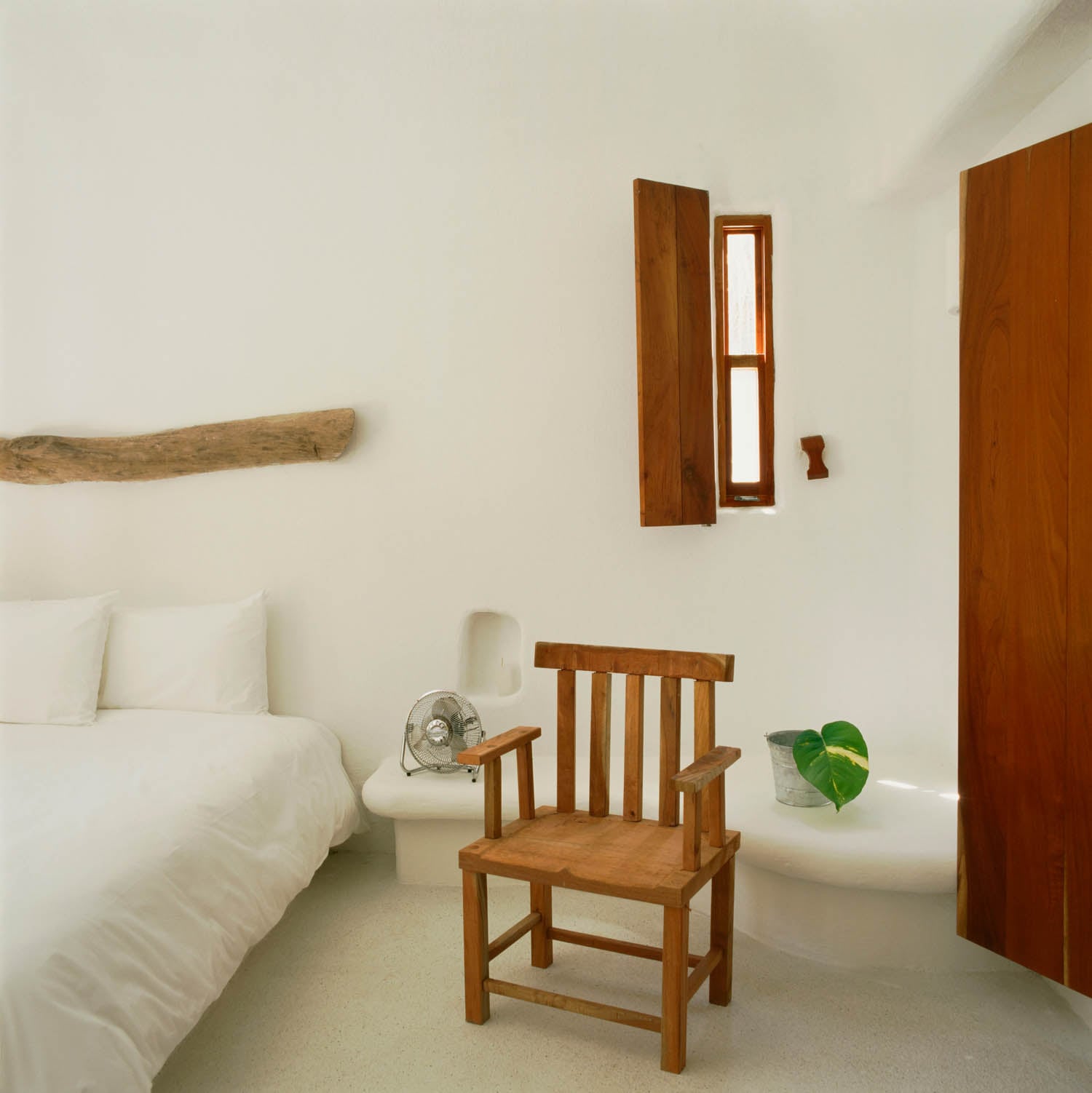 Minimalist bedroom with white walls, a wooden chair, a bed with white linens, a small fan, a large leaf in a glass vase, and wooden window shutters.