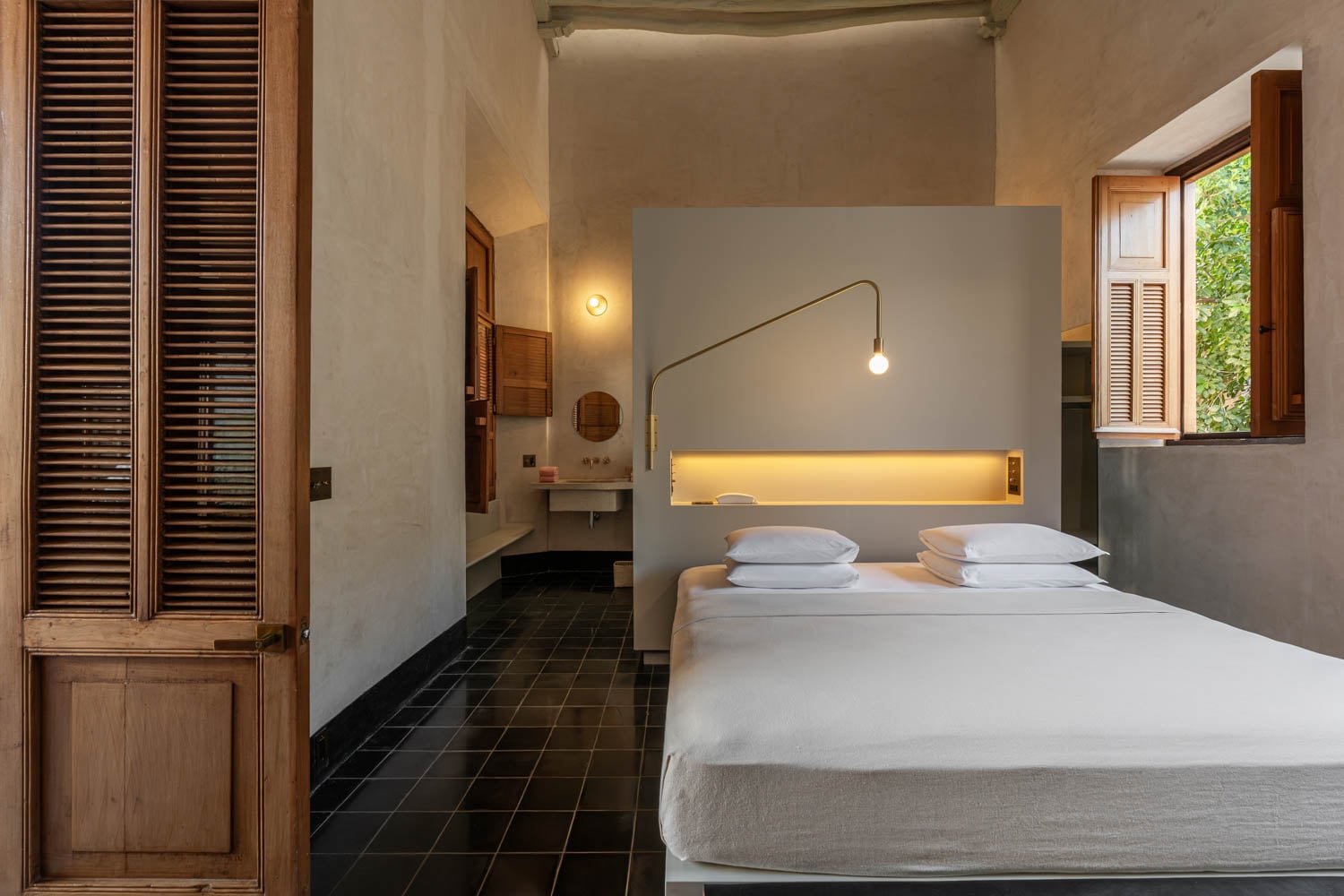 A minimalist bedroom with black tile floor, white bed, modern lighting, wooden shutters, and an open window letting in natural light.