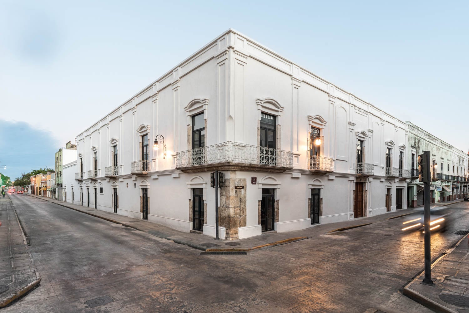 A large, two-story white colonial-style building stands on a street corner with balconies and ornate details, illuminated by streetlights at dusk.