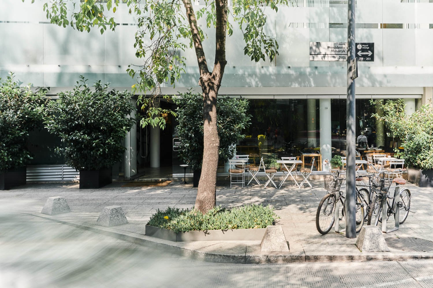 Street view of a modern café with outdoor seating, several tables and chairs, potted plants, trees, and two bicycles parked at a bike rack.