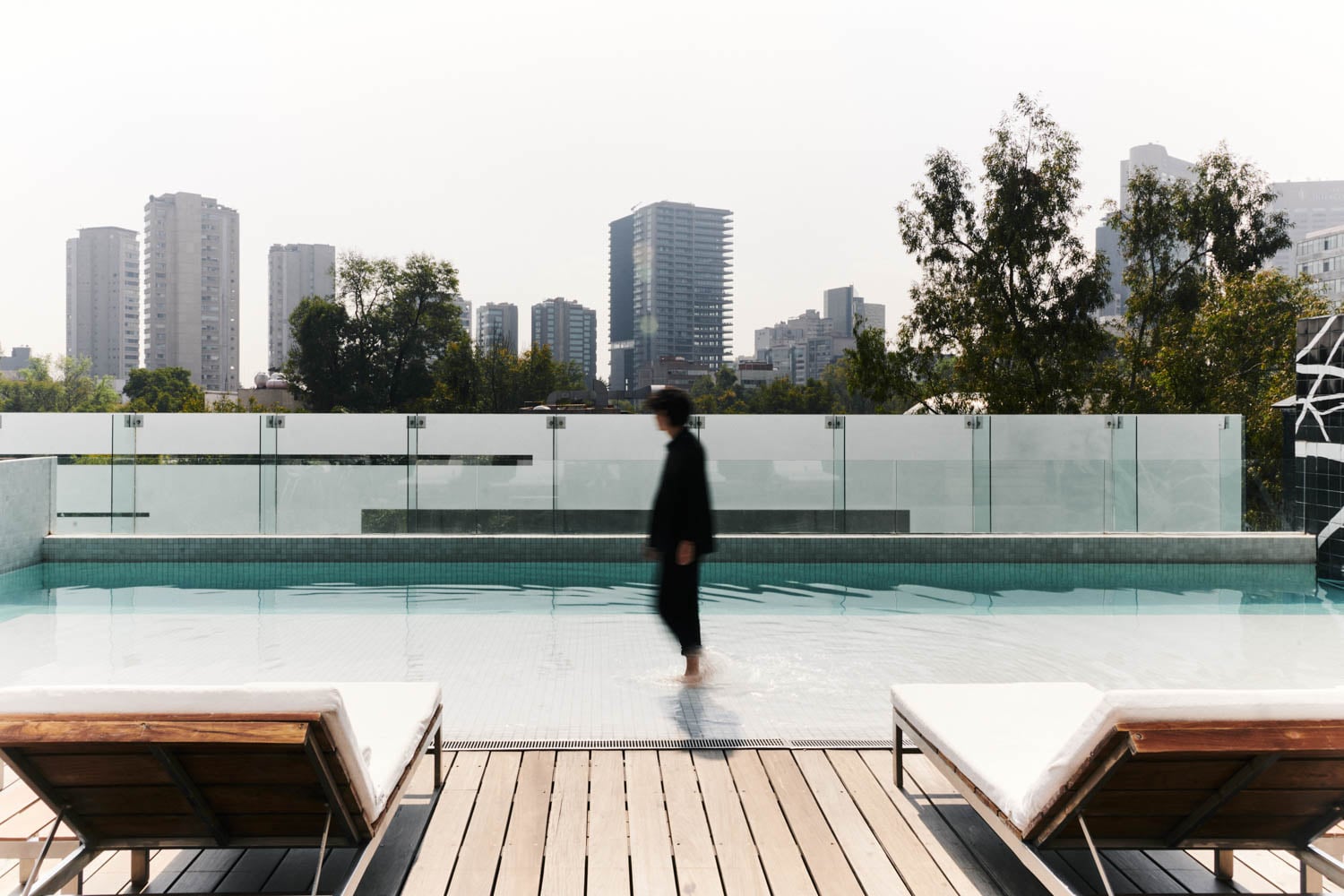 A person walks along the edge of a rooftop pool with city buildings and trees in the background; two empty lounge chairs are in the foreground.