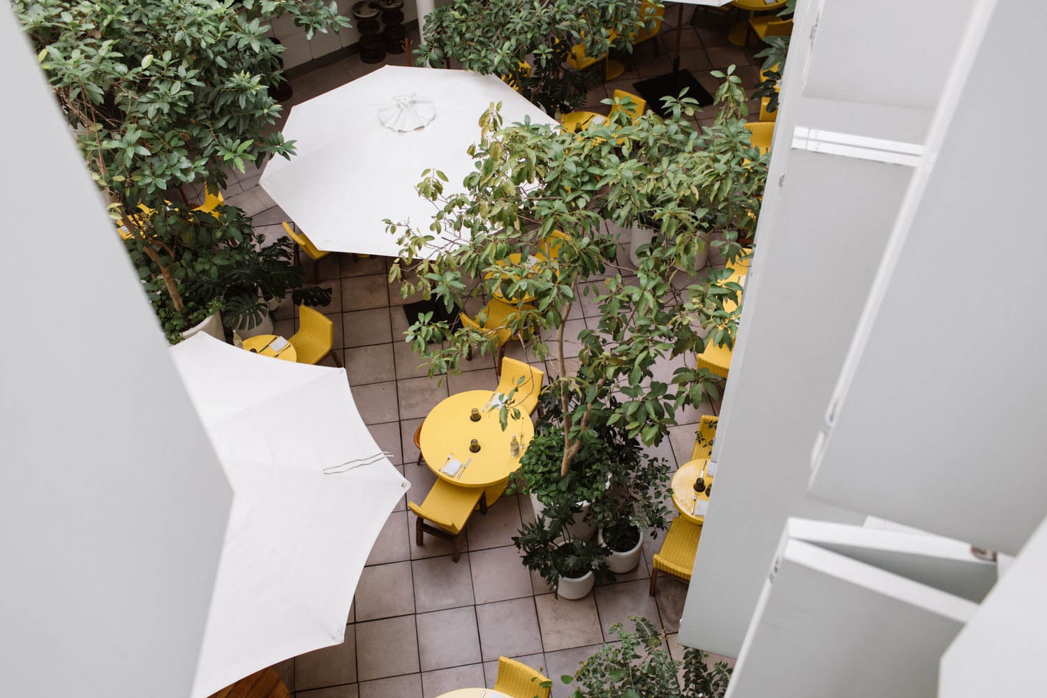 Aerial view of an outdoor cafe with yellow tables and chairs, large white umbrellas, and potted green plants arranged on a tiled floor.
