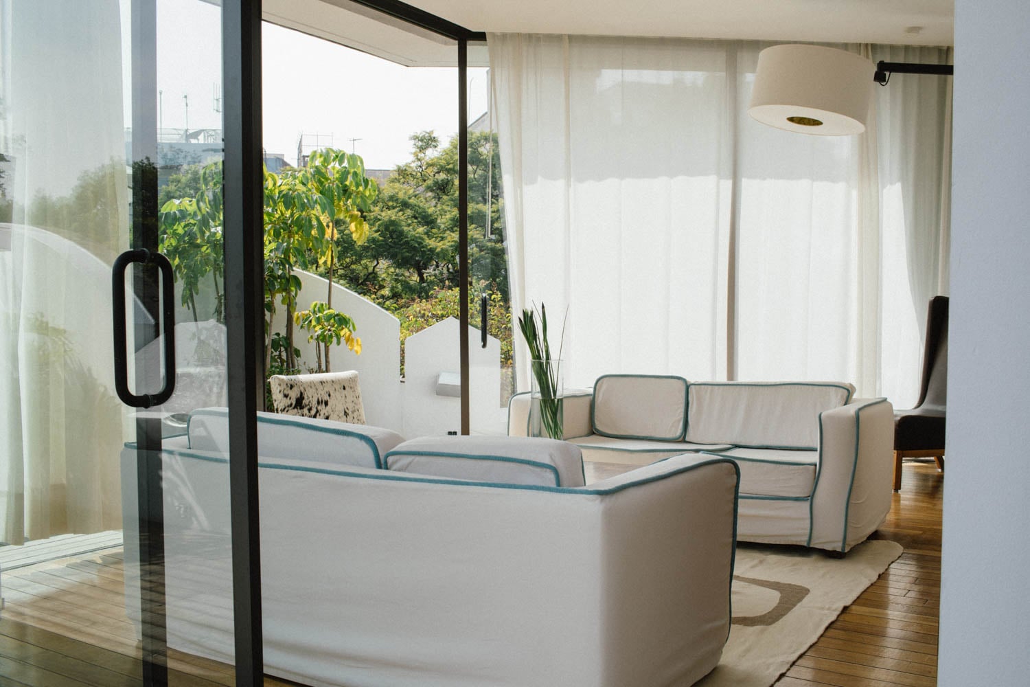 Modern living room with two white sofas, a floor lamp, and large windows overlooking greenery; sunlight filters through sheer curtains.