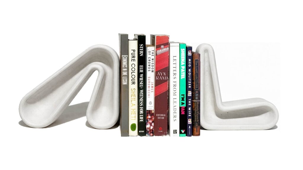 Seven books stand upright between two modern, white, sculptural bookends shaped like bent tubes against a white background.