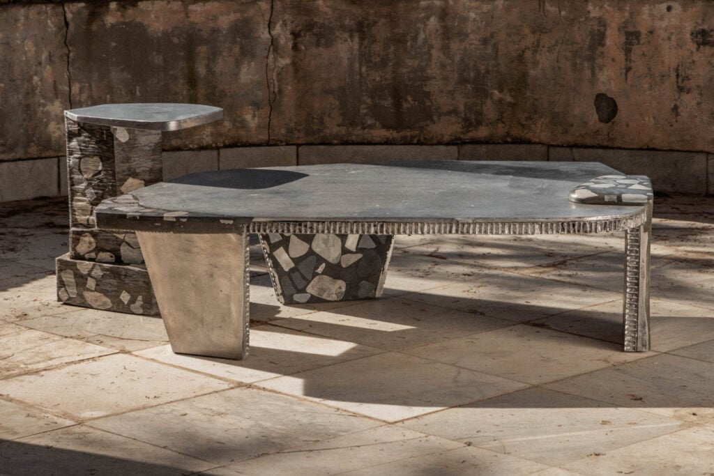 Abstract stone and metal table with geometric design and matching side table, set on a tiled floor against a weathered wall.