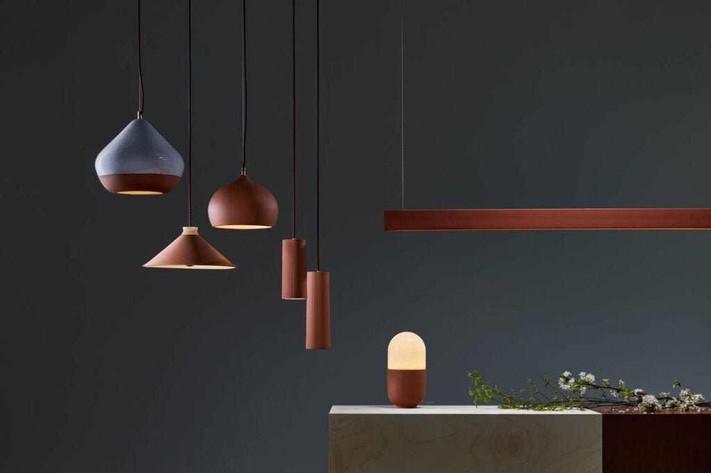 Five modern pendant and table lamps in muted colors hang or sit against a dark background, with a small branch of white flowers on a light wood surface.