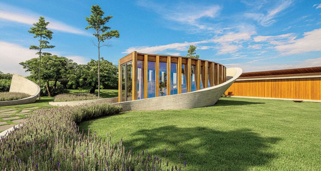 A modern building with curved concrete and wood elements sits on a manicured lawn with lavender plants and trees under a clear blue sky.