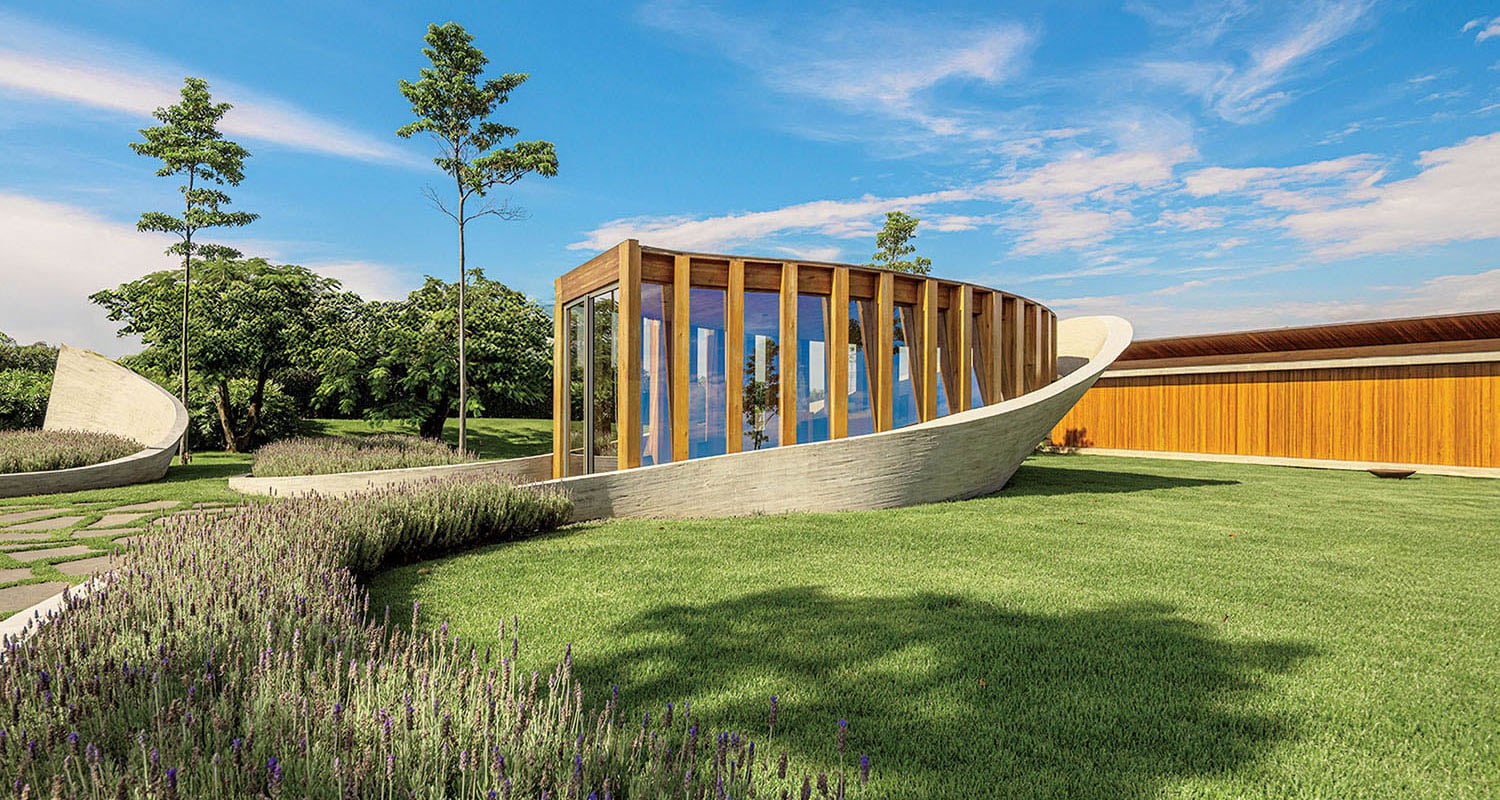 A modern building with curved concrete and wood elements sits on a manicured lawn with lavender plants and trees under a clear blue sky.