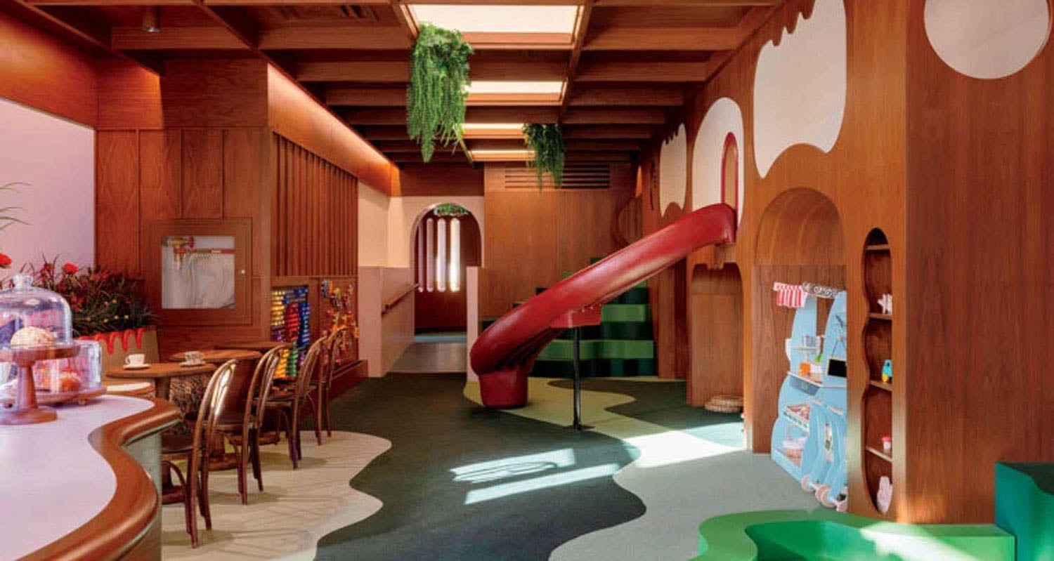 A children’s play area with wood-paneled walls, a red slide, tables and chairs, greenery, and built-in shelves with toys and books.