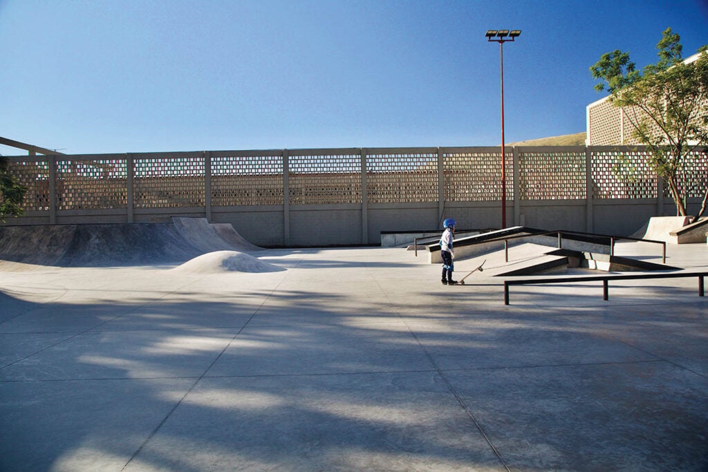 A child wearing a helmet stands with a scooter in an empty outdoor skate park with ramps, rails, and concrete structures under a clear sky.