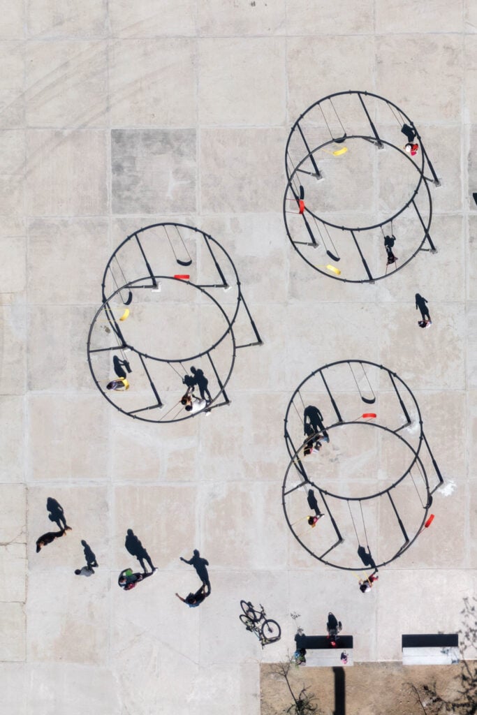 Aerial view of three circular swing sets with people using them and others walking nearby on a paved surface.