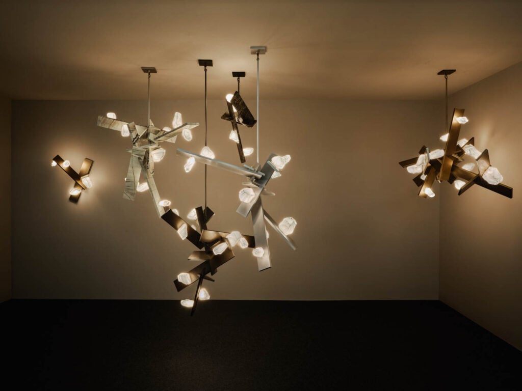 A room with three modern chandeliers featuring abstract, angular metal shapes and multiple exposed light bulbs, casting soft light on the walls.