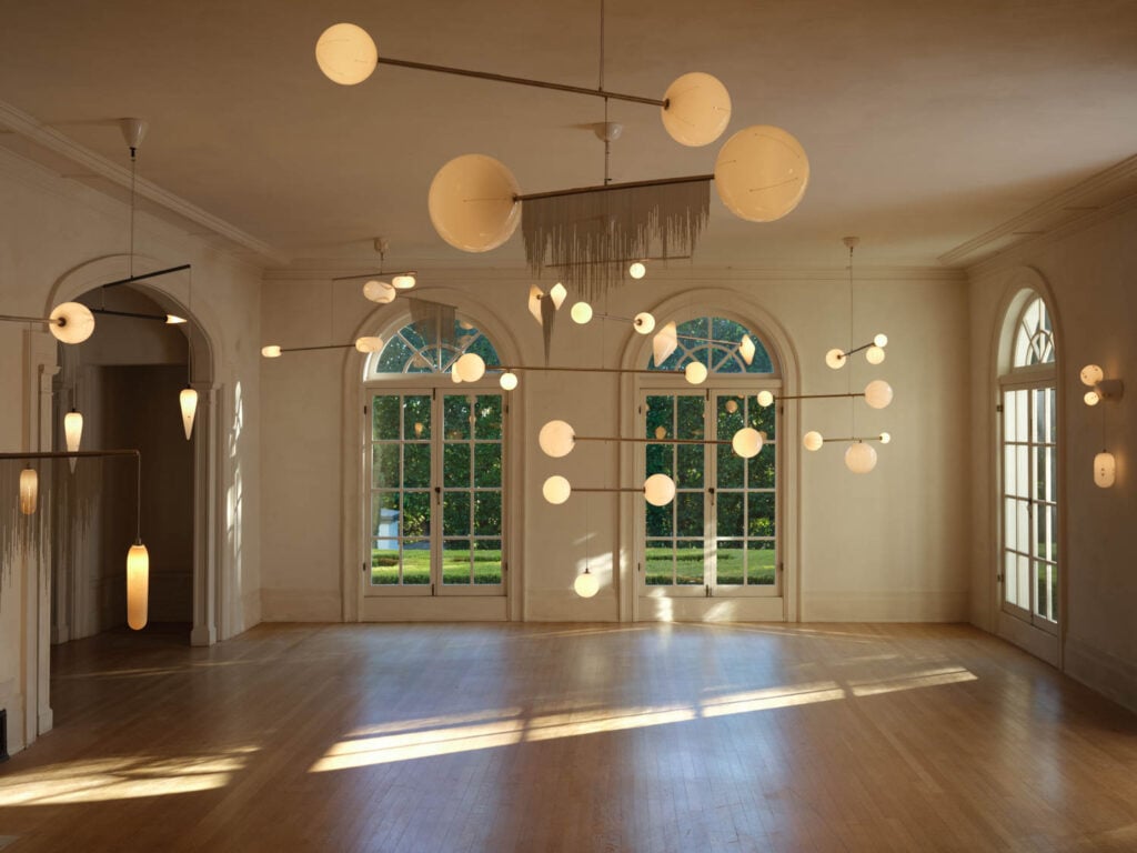 A spacious, sunlit room with large arched windows, wooden floors, and multiple modern pendant lights with round and elongated white globes hanging from the ceiling.