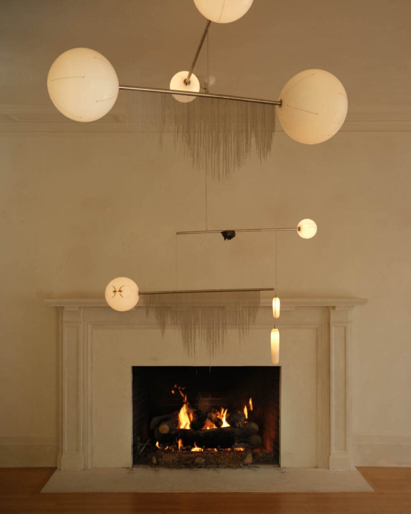 A modern chandelier with spherical lights hangs above a lit fireplace in a minimalist room with a white mantel and neutral walls.