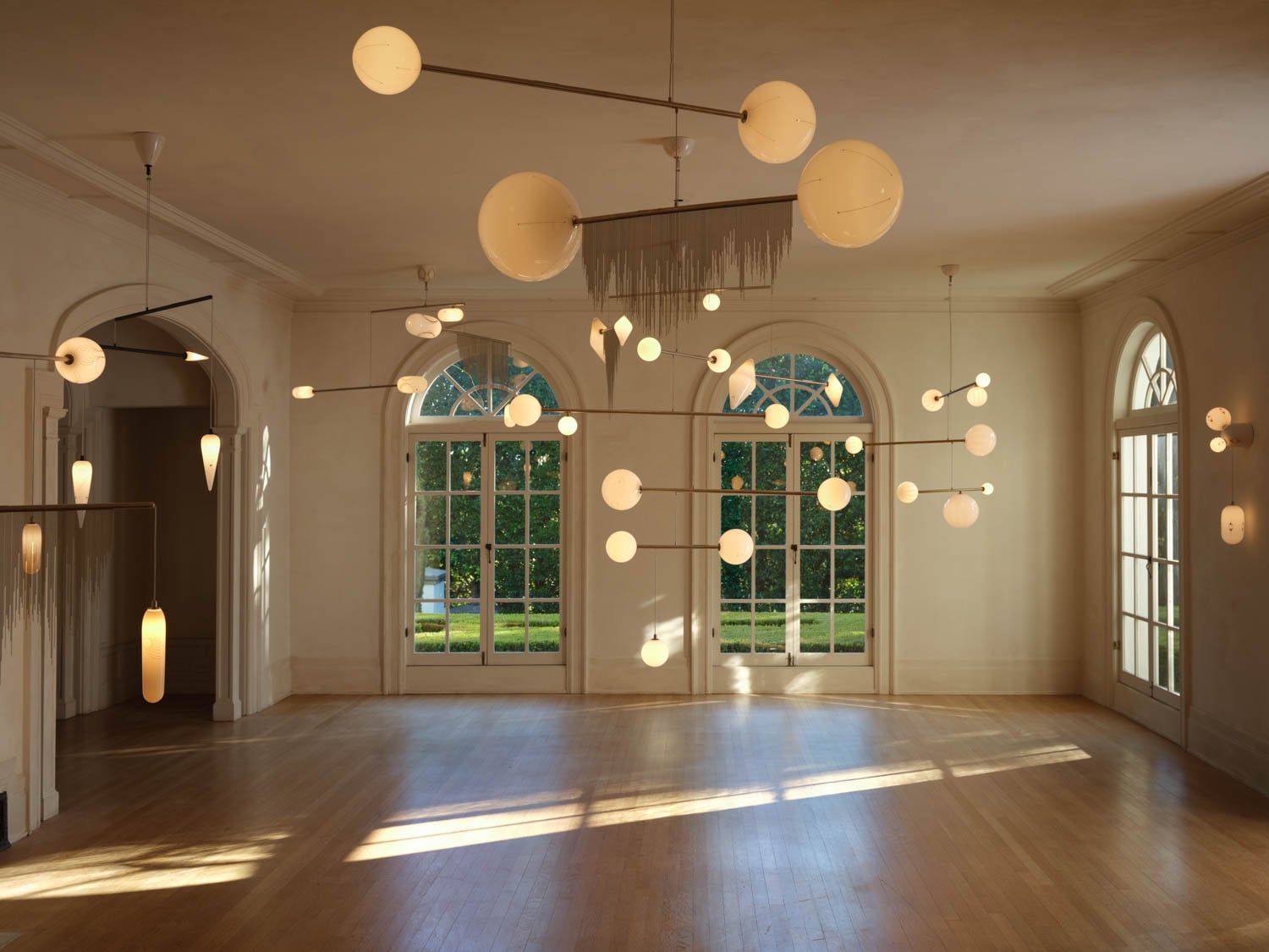 A spacious, sunlit room with large arched windows, wooden floors, and multiple modern pendant lights with round and elongated white globes hanging from the ceiling.