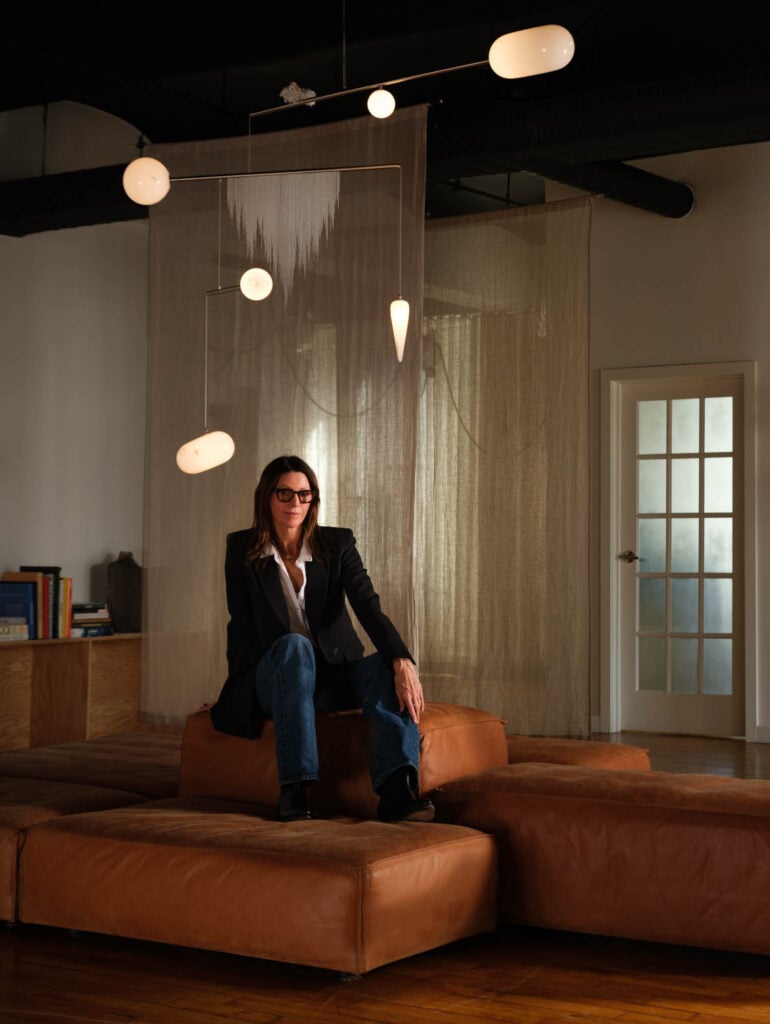 A woman in a black blazer, jeans, and sunglasses sits on a brown leather sectional in a modern room with a hanging light fixture and sheer curtain background.