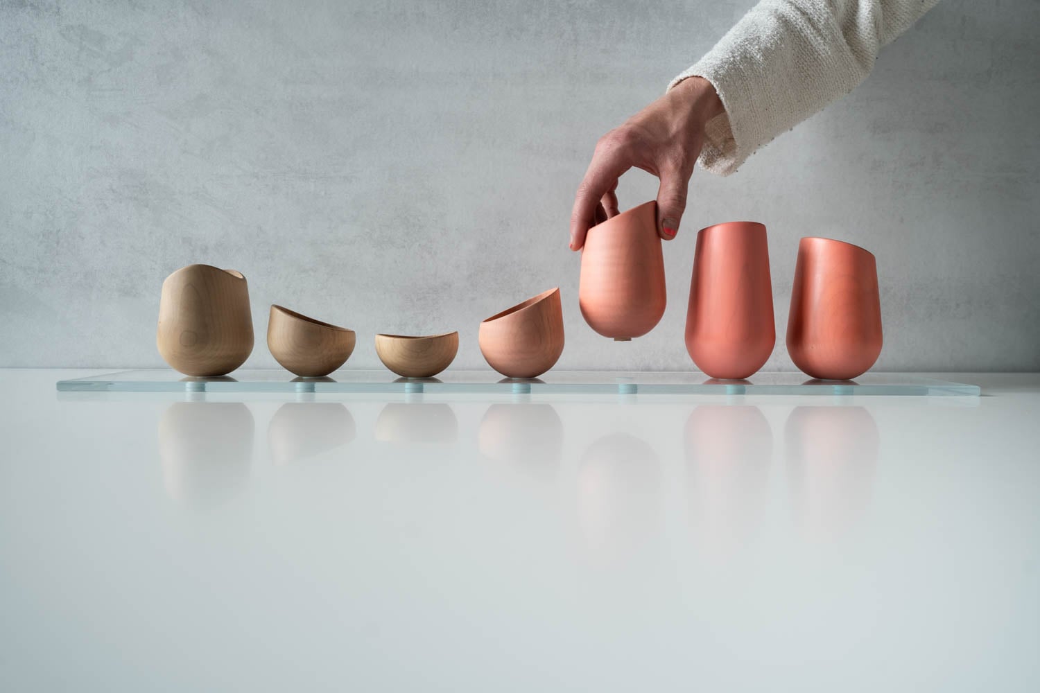 A hand picks up one of several modern, differently sized wooden and ceramic cups arranged in a row on a glass surface.