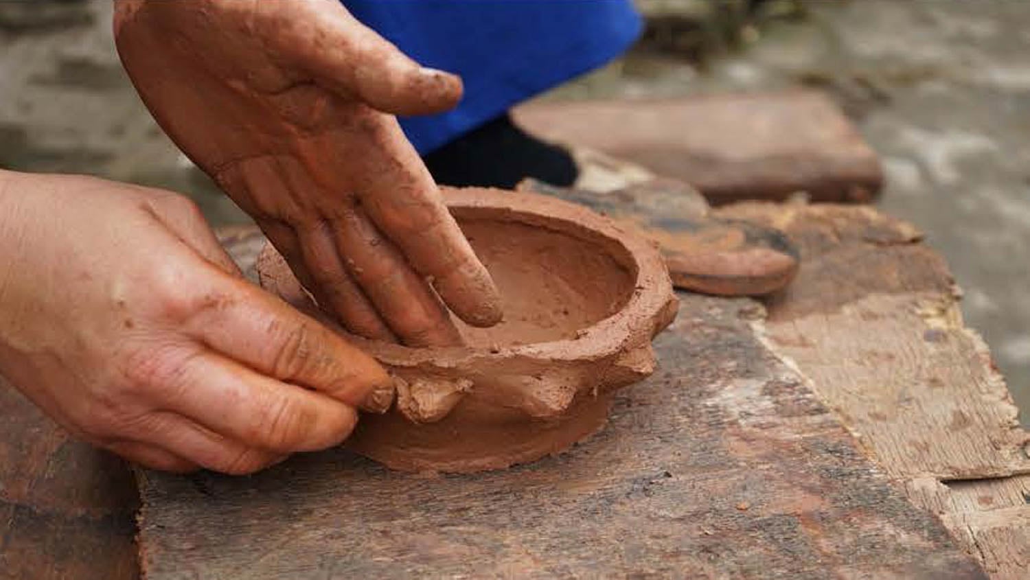 Hands shaping a small clay pot on a wooden surface, with clay smudges visible on the fingers and pot.