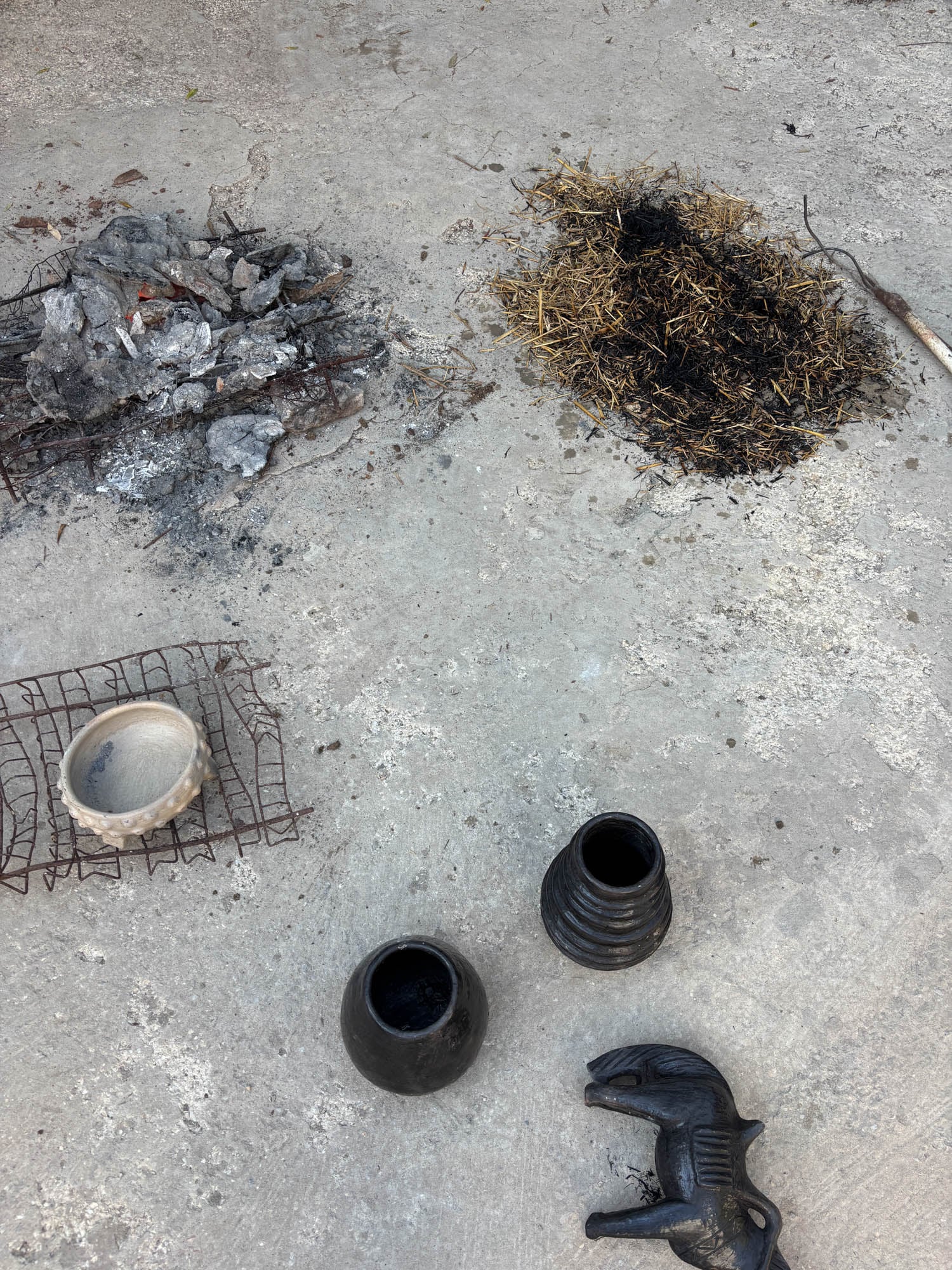 Ceramic vessels and a horse figurine sit on a concrete surface near piles of ash, straw, and a metal grate, suggesting recent firing or pottery-making activity.
