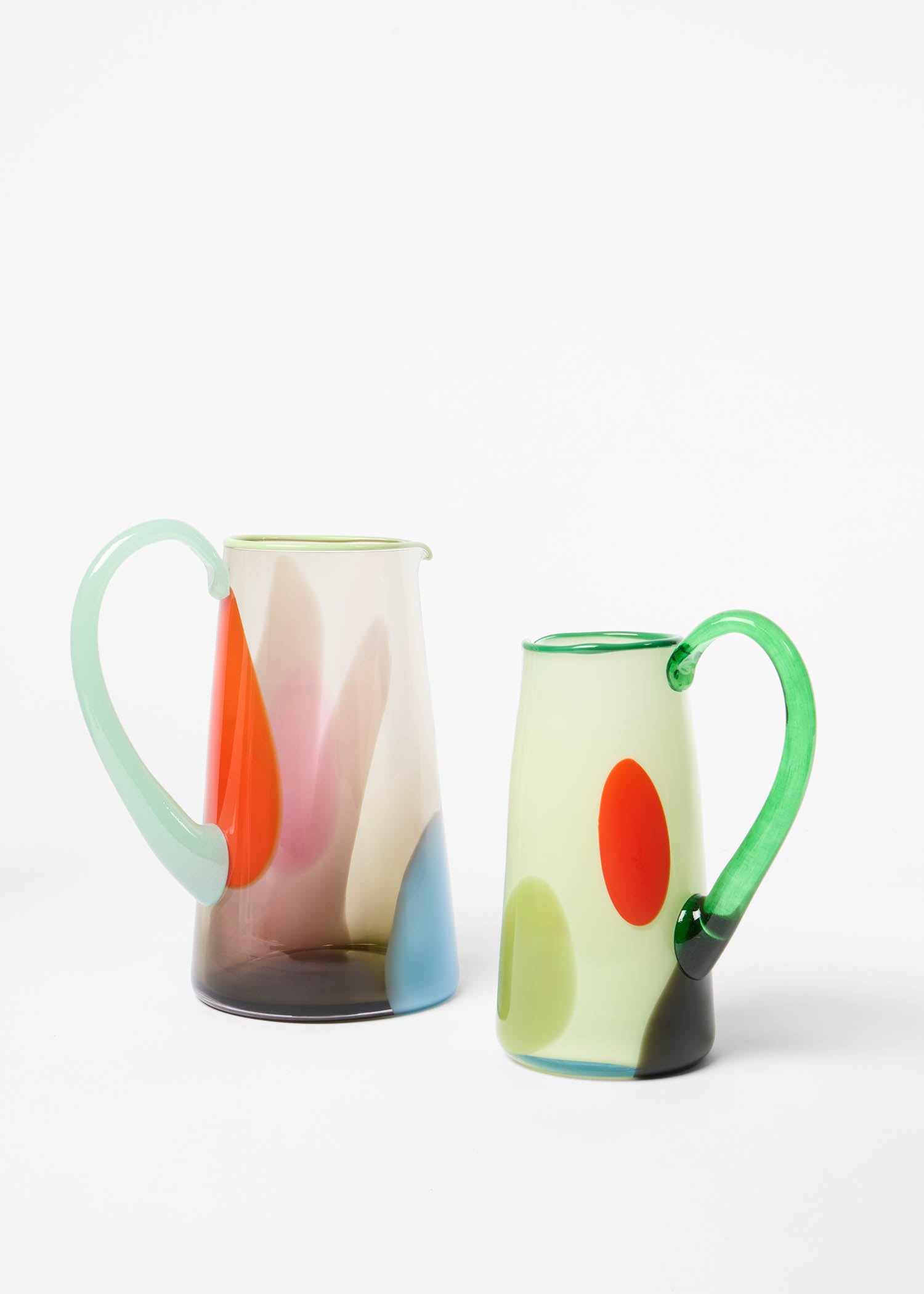 Two glass pitchers with abstract, colorful patterns and green handles, set against a plain white background.