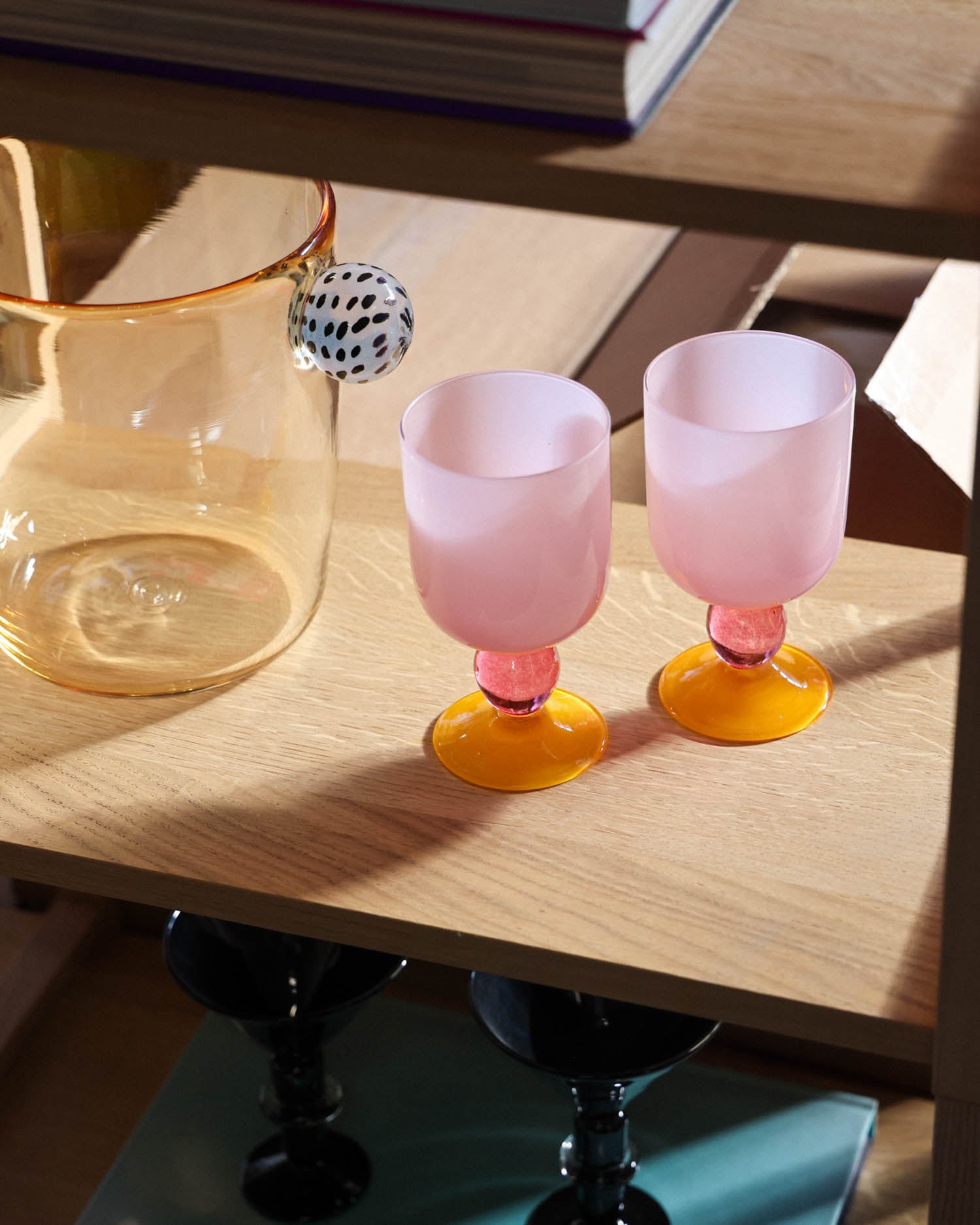 Two pink goblets with orange bases sit on a wooden shelf next to a transparent pitcher with an orange tint and a black-and-white patterned handle.