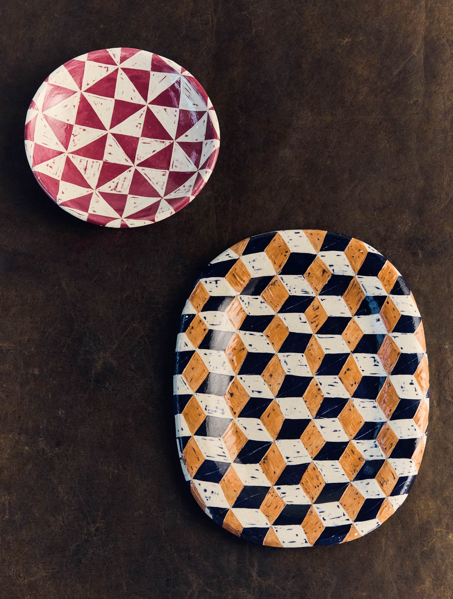 A round plate with a red and white geometric pattern and an oval platter with a yellow, black, and white 3D cube pattern on a dark surface.