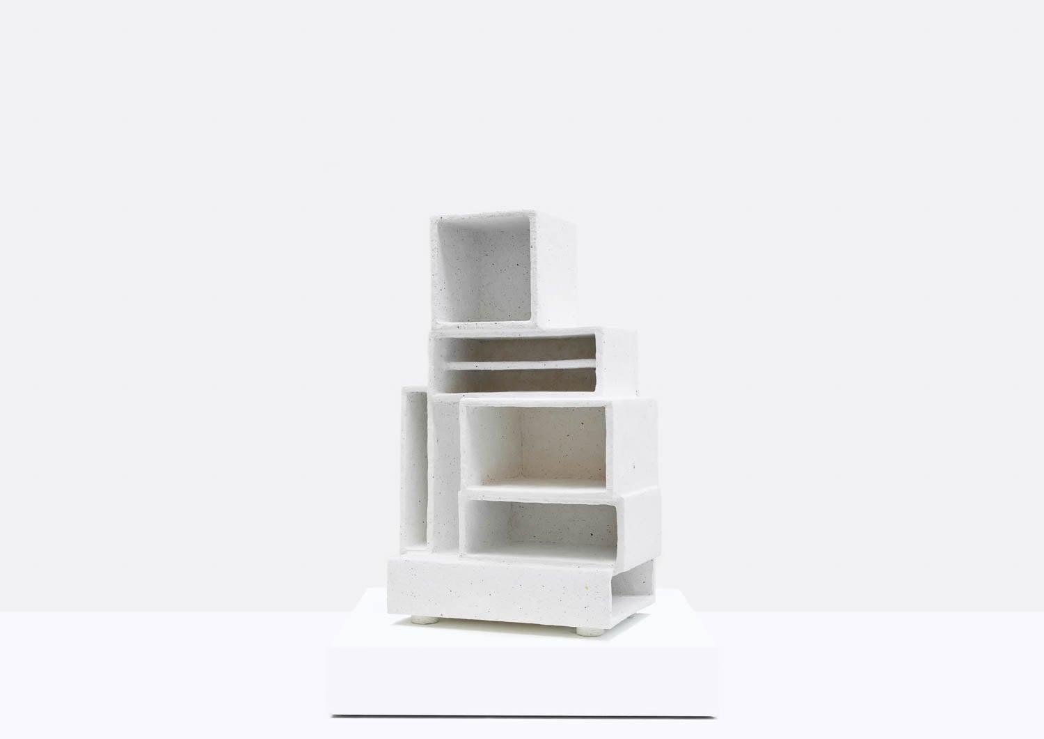 A white, abstract sculpture composed of stacked, rectangular open boxes arranged unevenly on top of each other, displayed on a white pedestal against a plain background.