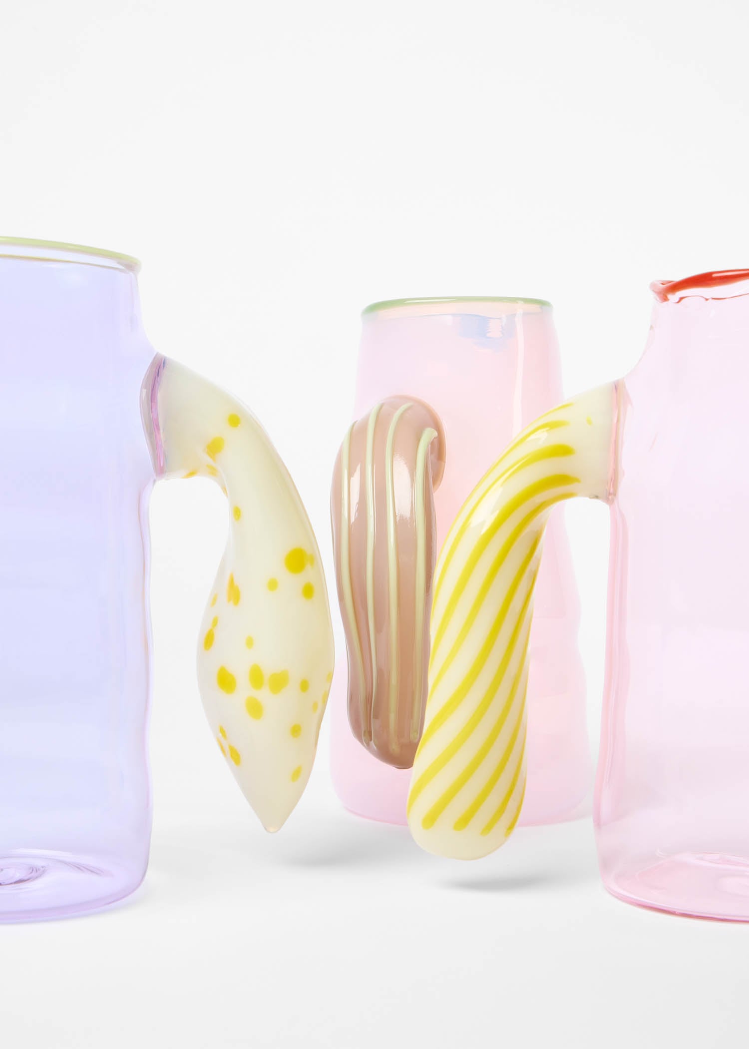 Three colorful glass mugs with uniquely shaped and patterned handles in yellow polka dots, beige stripes, and yellow stripes are arranged against a white background.
