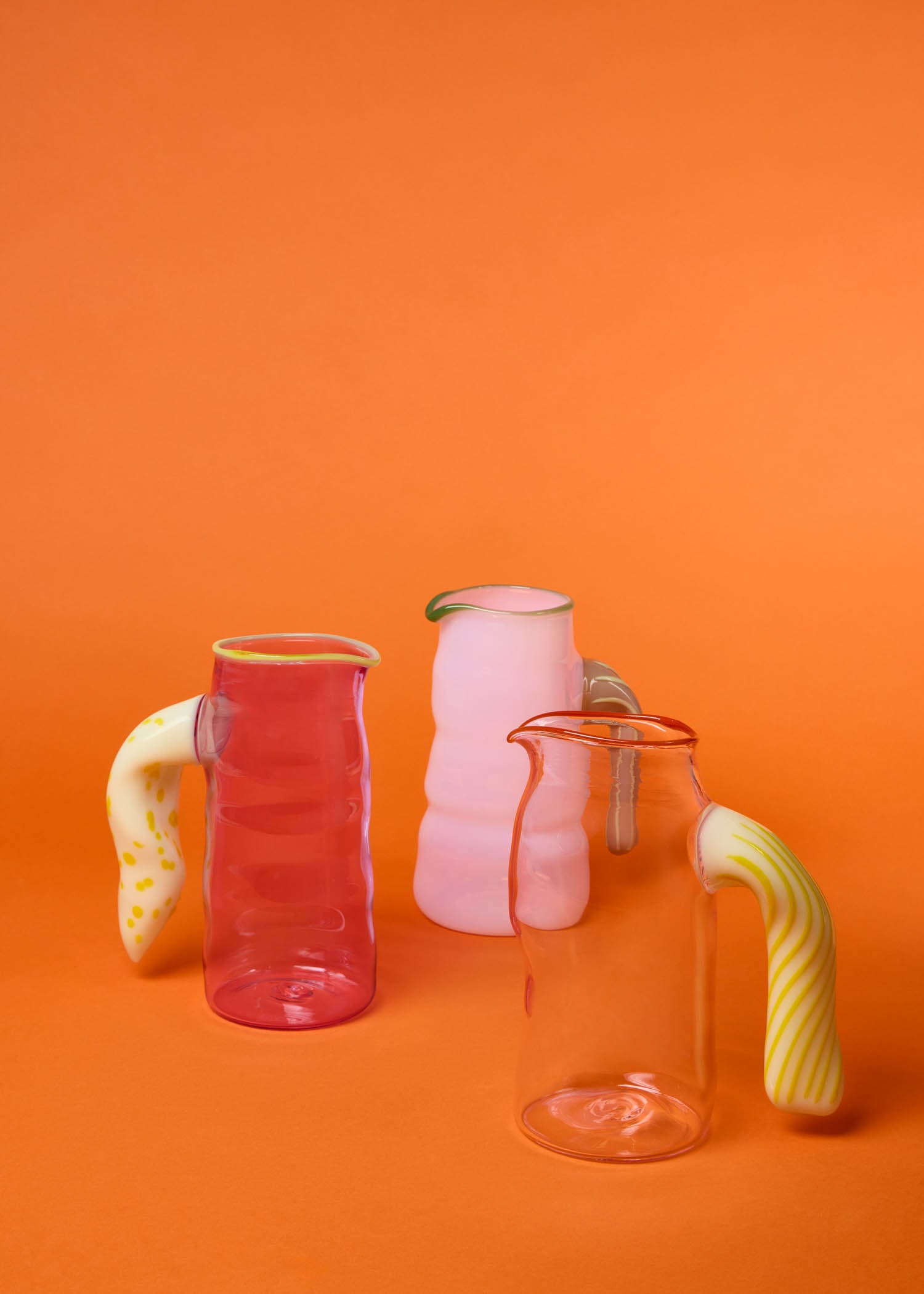 Three glass pitchers with wavy handles—one pink, one clear, one red—stand on an orange background. The handles are yellow, white, or marbled in color.