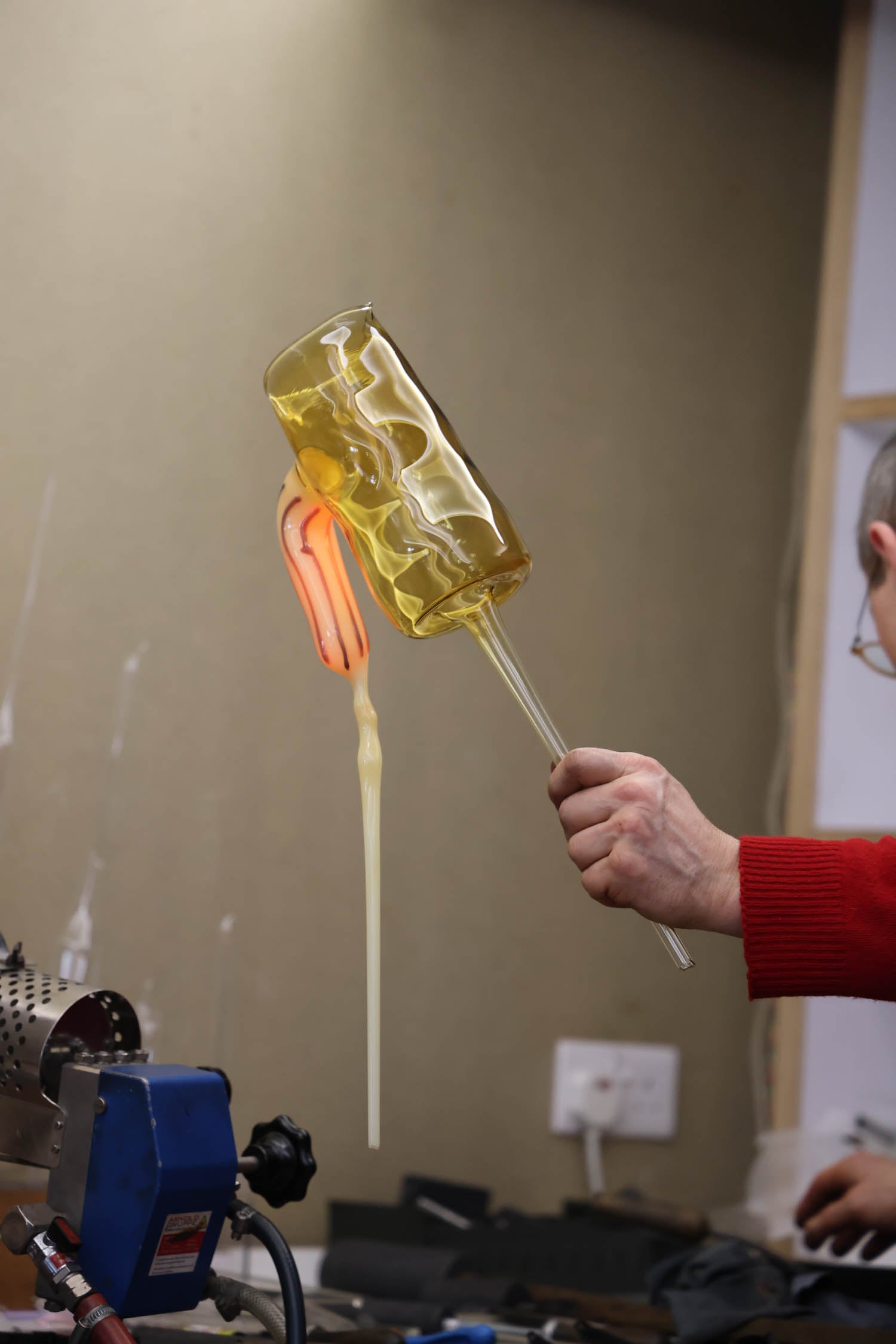 A person in a red sweater holds a glass-blowing pipe with molten glass, forming a yellowish glass object inside a workshop.