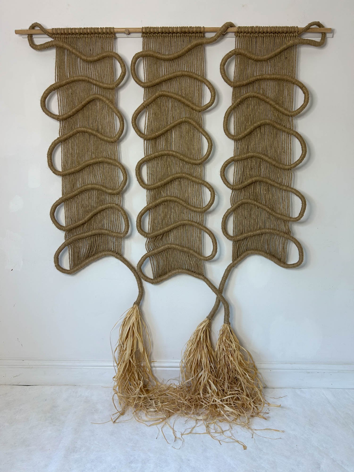 A wall hanging of three vertical woven ropes with looping patterns, ending in fringe-like strands at the bottom, displayed against a white wall.
