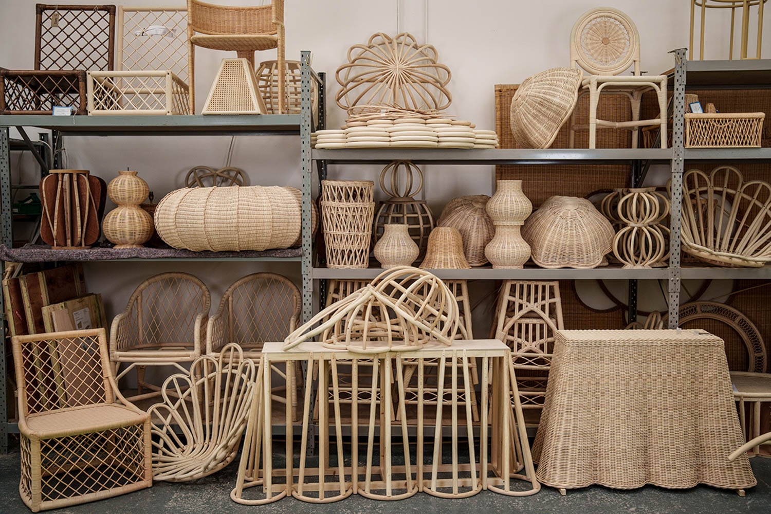 Shelves display various wicker and rattan furniture and decor items, including chairs, baskets, lampshades, stools, and tables in a storage or workshop setting.