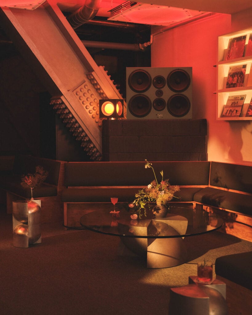 A dimly lit lounge with a sectional sofa, glass coffee table with a floral arrangement, drinks, large speakers, and framed photos on the wall. Warm orange lighting fills the space.
