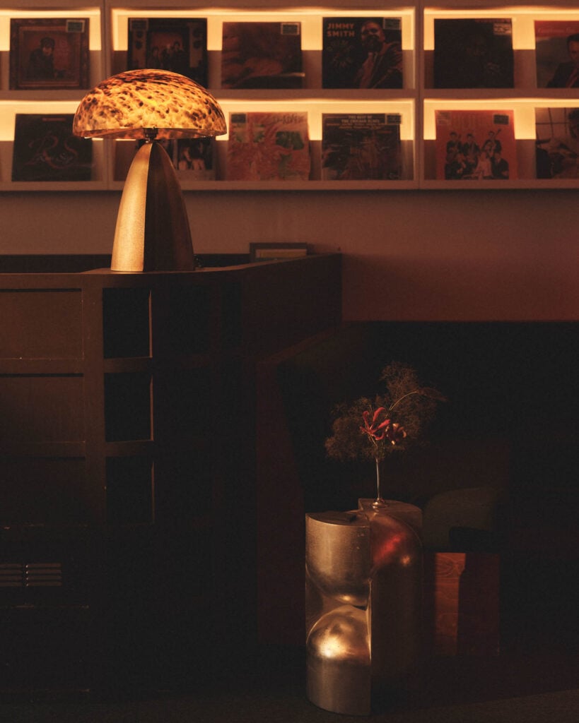 A warm-lit room with a mushroom-shaped lamp on a dark wooden shelf, a vase with dried flowers, and a wall display of vinyl records in the background.