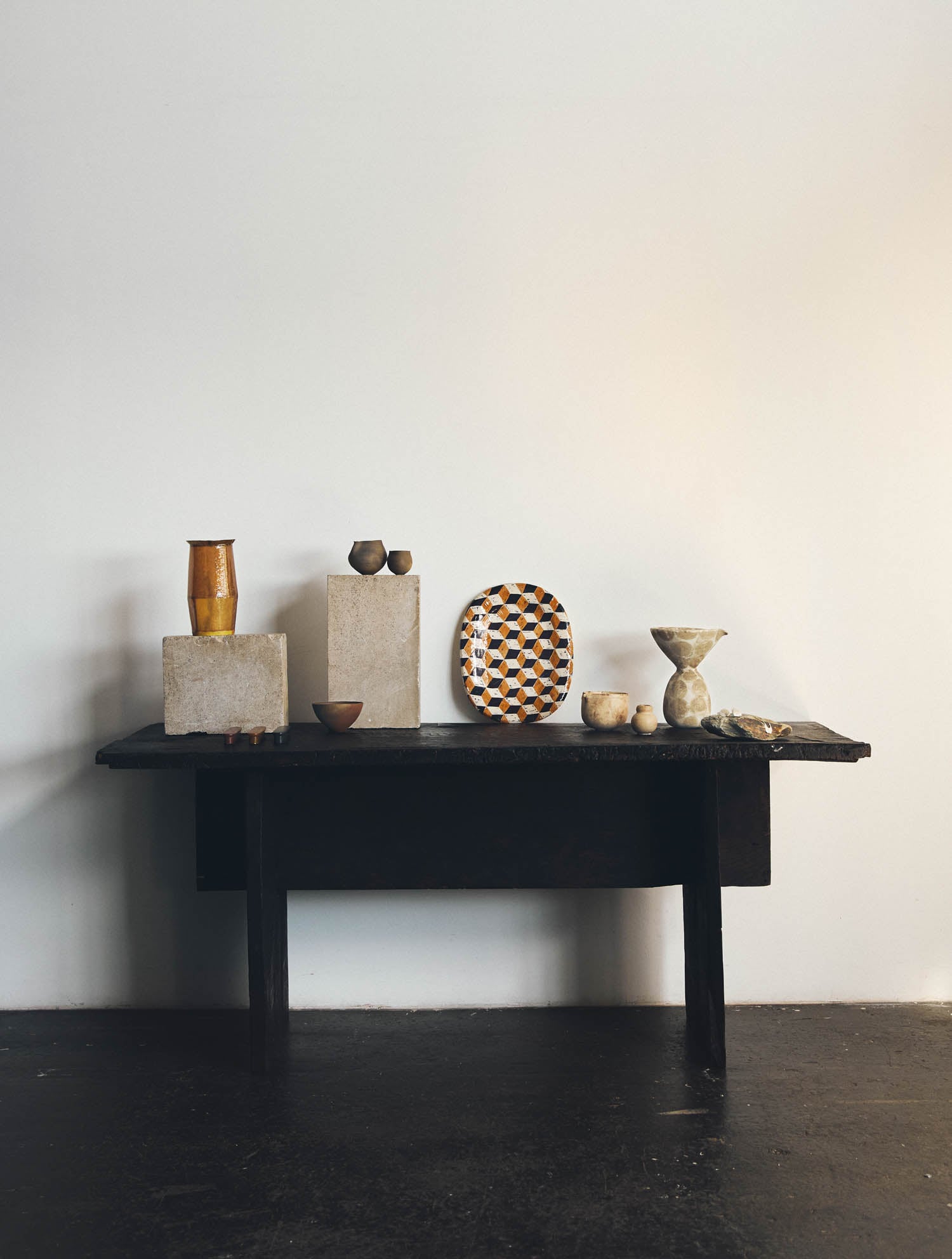 A dark wooden table against a white wall displays ceramic vases, bowls, a decorative plate, and small sculptural objects arranged on and around stone blocks.