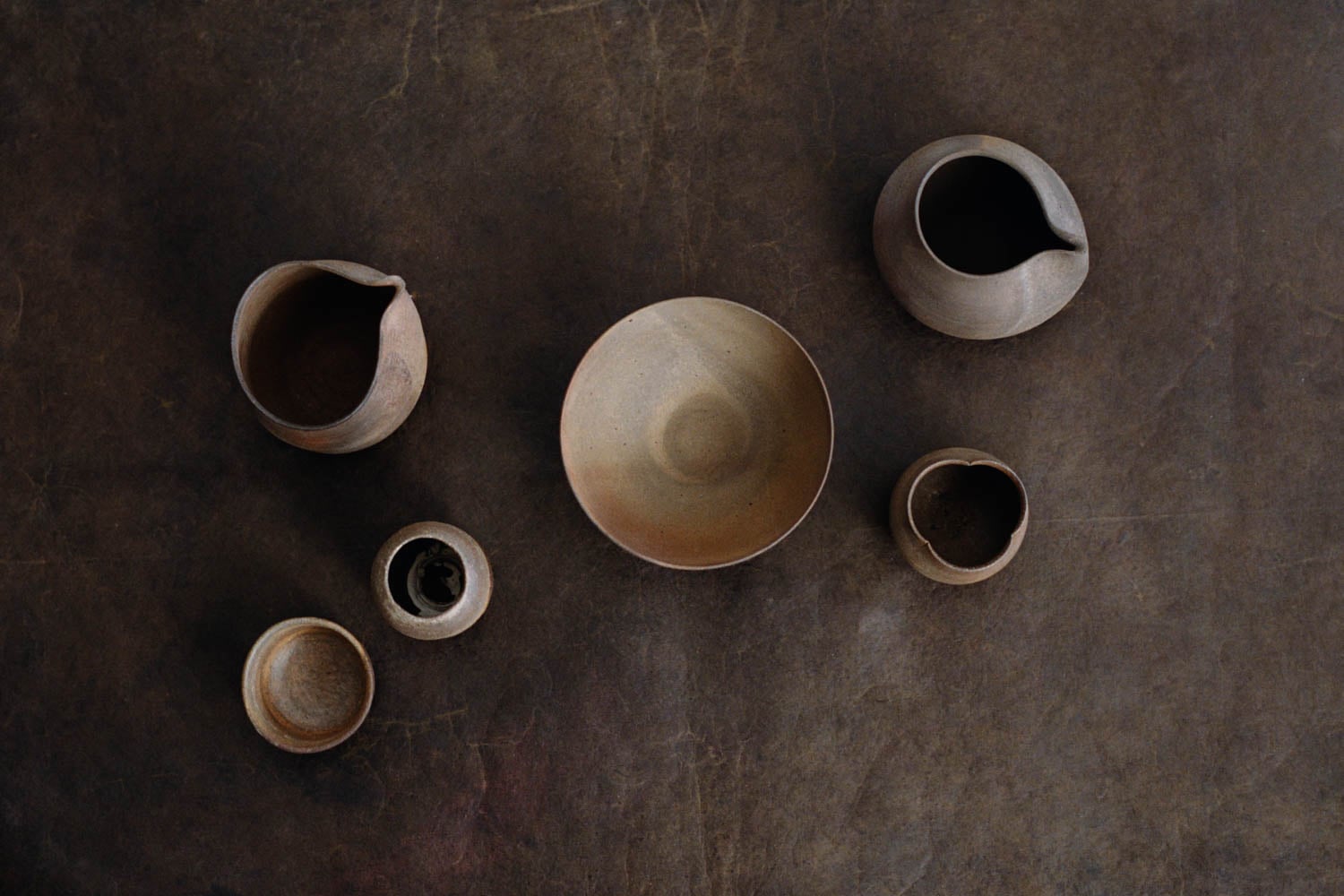 Six handcrafted clay vessels of varying shapes and sizes are arranged on a dark, textured surface viewed from above.