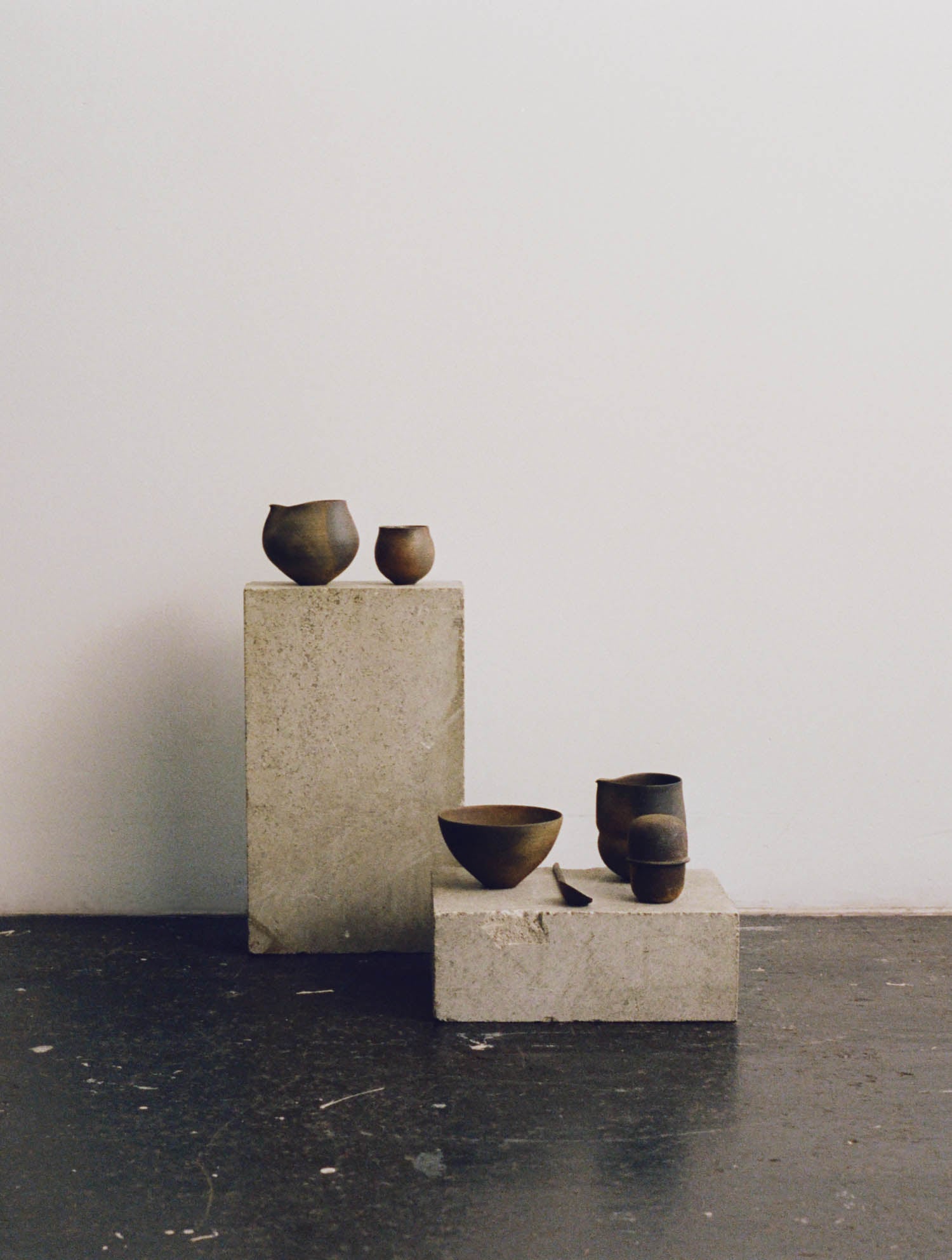 Five small, dark-toned ceramic vessels are displayed on two rectangular concrete blocks against a plain white wall and a black floor.