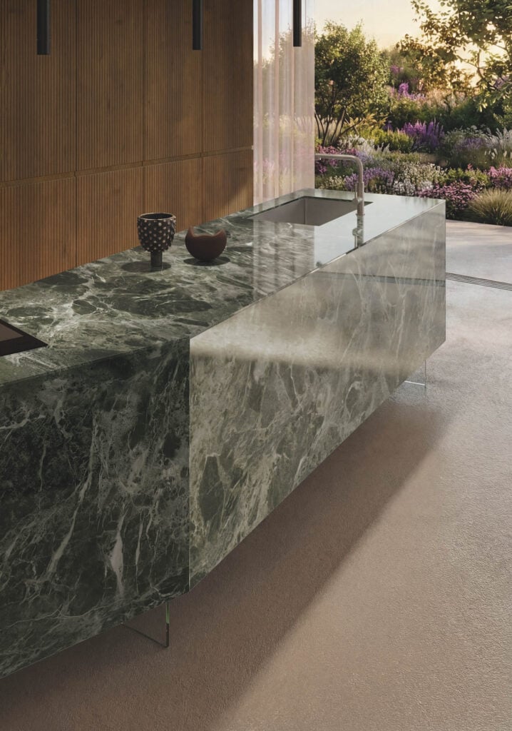 Modern kitchen with a large marble island, built-in sink, minimalist decor, and a view of a landscaped garden through floor-to-ceiling windows.