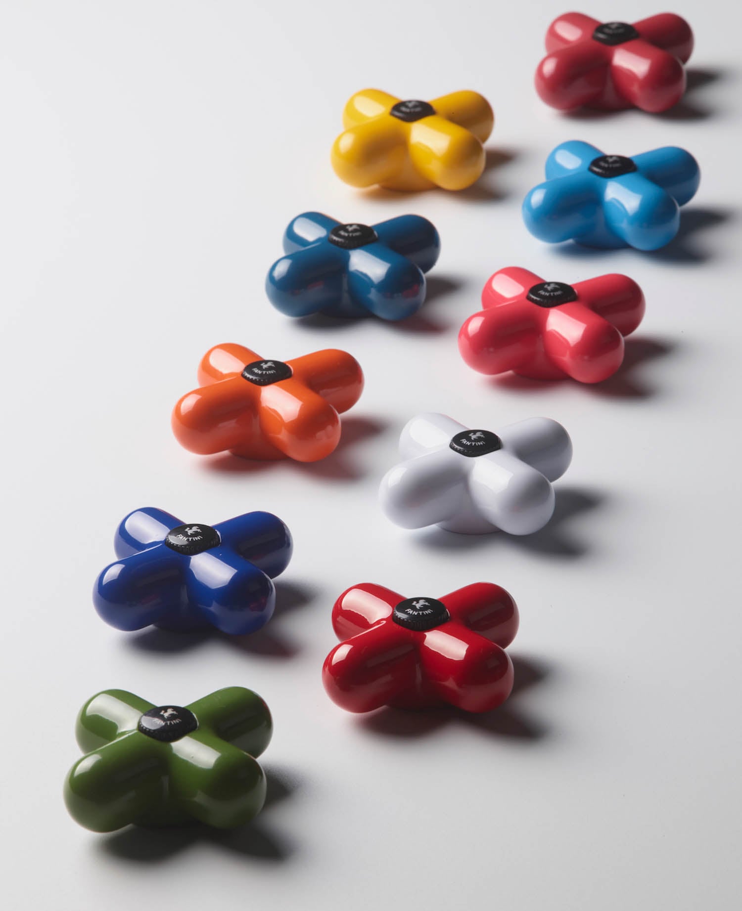Ten colorful, glossy, four-lobed objects are arranged in a loose diagonal line on a white surface. Each object has a black label with a white symbol in the center.
