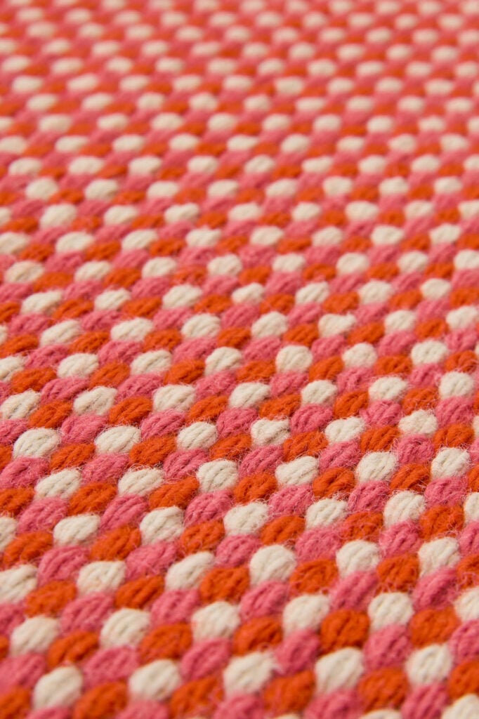 Close-up of a textured fabric with a repeating pattern of pink, orange, and white woven yarn.