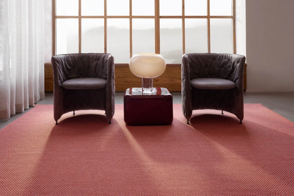 Two dark leather armchairs face forward, separated by a small table with a white modern lamp, all set on a textured red rug in front of a large window with sheer curtains.