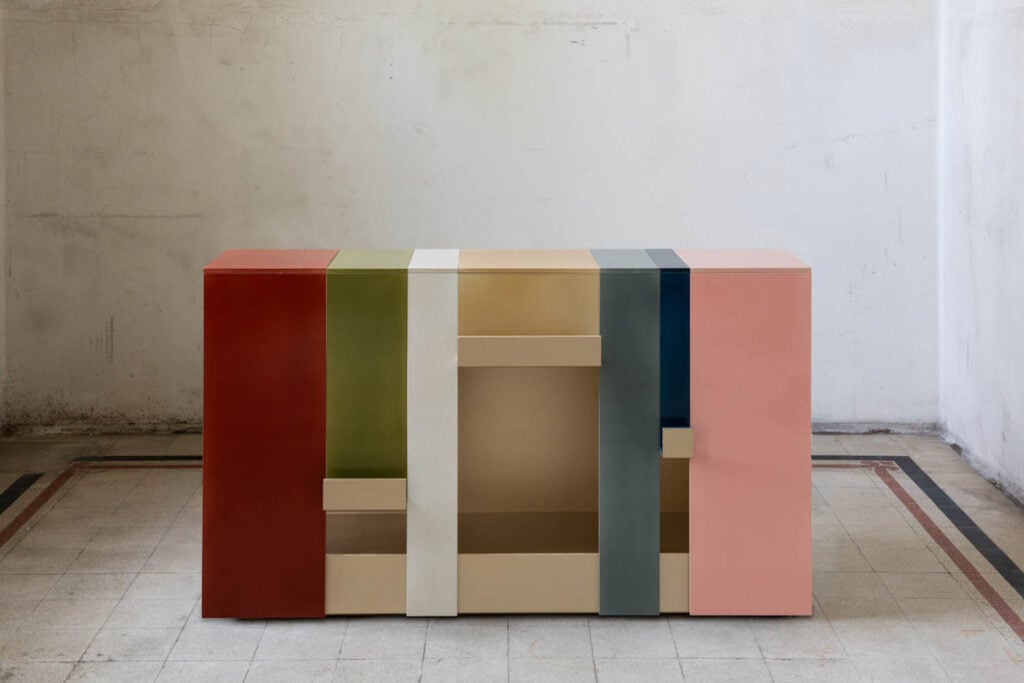 A modern sideboard with vertical color blocks in red, green, white, beige, gold, blue, and pink, positioned against a plain, textured wall and tiled floor.