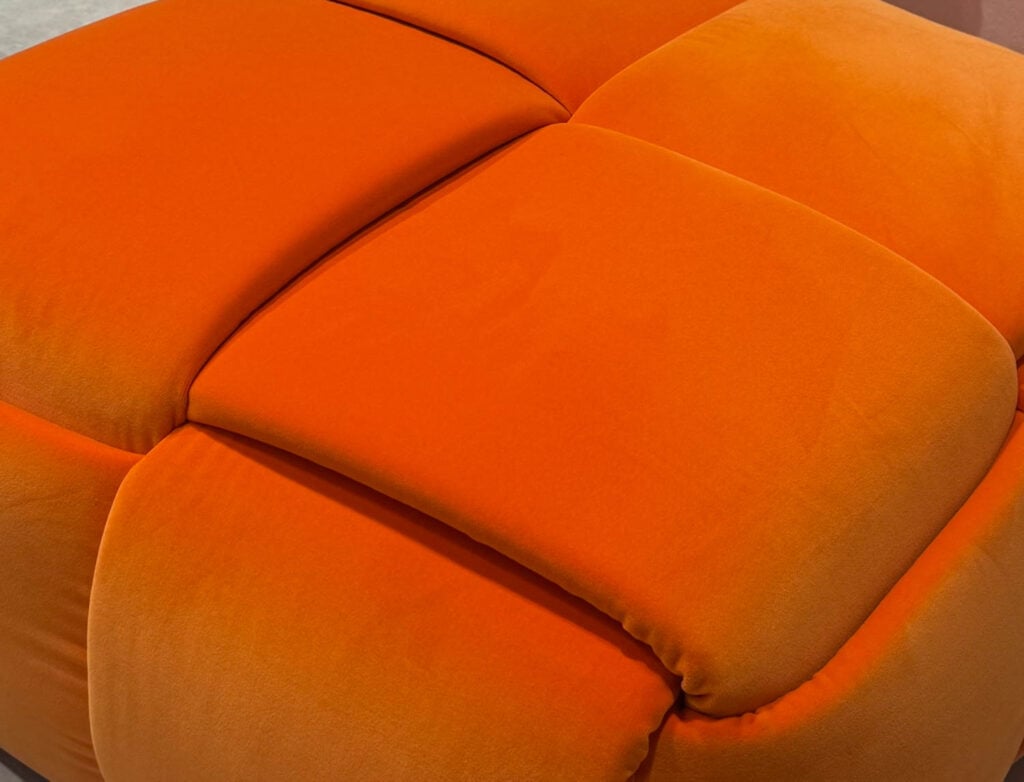 Close-up view of an orange, tufted, fabric ottoman with soft, cushioned squares and visible stitching.
